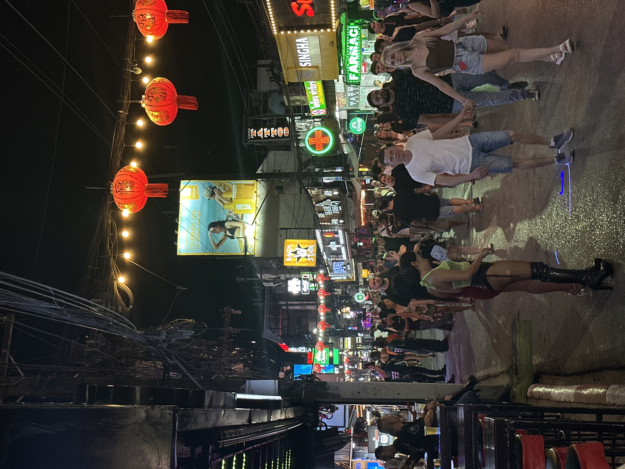 The scene depicts a bustling nighttime street in Patong, Thailand, characterized by a high concentration of commercial activity and pedestrian traffic. Overhead, an intricate network of tangled electrical wires is visible, strung between poles, alongside decorative string lights featuring bare bulbs and three prominent red Chinese lanterns with gold script and tassels.
The street is densely populated with a diverse crowd of pedestrians walking in both directions. In the foreground, slightly right of center, a Caucasian male in a white t-shirt and denim shorts is seen holding hands with a Caucasian female wearing a brown strap top and denim shorts, both walking forward. To the left, a woman in a neon green halter top, black fishnet stockings, and high-heeled black boots stands near the curb. Further left, several individuals are seated at outdoor bar tables with red stools, one person holding a glass containing a light-colored beverage.
The surrounding buildings are illuminated by numerous bright neon signs in various colors. Notable signs include "SINGHA," "Tattoo," a green cross sign indicating "City Pharmacy" and "FARMA Pharmacy," and a "ROCK STAR" sign. A large billboard on the right features a woman in a bikini, advertising "Celebrate Life." The ground is an asphalt surface, appearing damp in places, reflecting some of the artificial light. The atmosphere is vibrant and highly illuminated by artificial sources against the dark night sky.