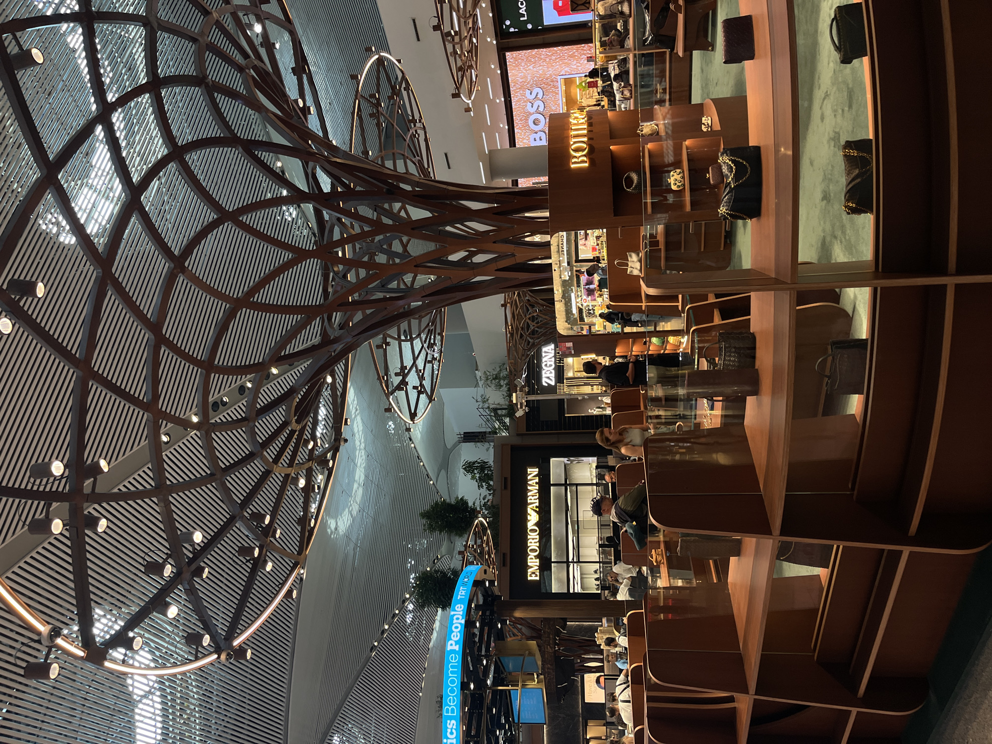 This is an indoor scene at Istanbul Airport, located in Arnavutköy, Turkey. The image captures a luxury retail area with prominent brand storefronts and display units. In the foreground, there are modern wooden shelving units showcasing various designer handbags, with a prominent black quilted handbag with gold chain detail displayed on the middle shelf. Behind these shelves, there are more shops visible, including "EMPORIO ARMANI," "ZEGNA," "BOSS," and "BOTTEGA VENETA." The ceiling features an intricate, tree-like metal structure with integrated lighting, creating a unique architectural element. Several people are present, some browsing in the shops and others seated at tables in the background, suggesting a bustling commercial environment. The overall aesthetic is sleek and contemporary, typical of a high-end international airport's retail offerings.