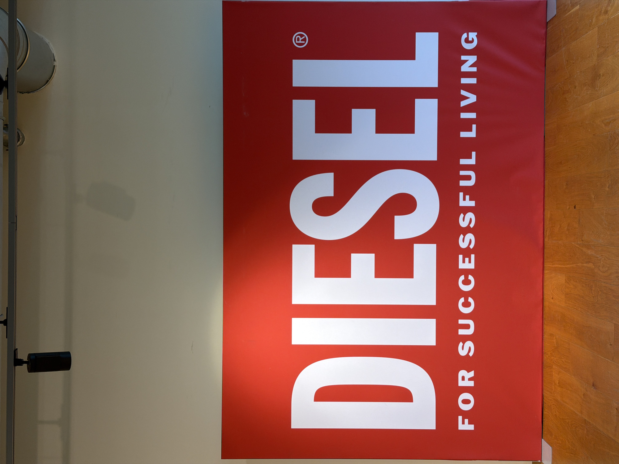 This image captures a large, vertically oriented red banner on the right side of the frame, emblazoned with the "DIESEL" brand logo. The banner features the word "DIESEL" in prominent white capital letters, reading from bottom to top in the current orientation of the image. A registered trademark symbol (®) is visible above the 'E' of "DIESEL". To the right of the main brand name, the tagline "FOR SUCCESSFUL LIVING" is written in smaller white capital letters, also oriented vertically and reading from bottom to top.
The left side of the frame is dominated by a light-colored, possibly off-white, wall. Towards the upper left corner, several metallic, cylindrical objects, resembling light fixtures or speakers, are mounted on the wall. In the lower left, a black cylindrical object, possibly a security camera or another light fixture, extends from the wall. The bottom right corner of the image reveals a segment of a light brown wooden floor with visible planks.
The scene depicts an indoor setting, likely a commercial space such as a retail store, showroom, or exhibition area in Mirabella, Italy, given the prominent branding. The banner is well-lit, suggesting direct artificial lighting, which casts subtle shadows on the adjacent wall. There are no people visible, and no specific activity is taking place; the image focuses on the architectural elements and the brand display. The overall impression is one of modern, clean commercial design.