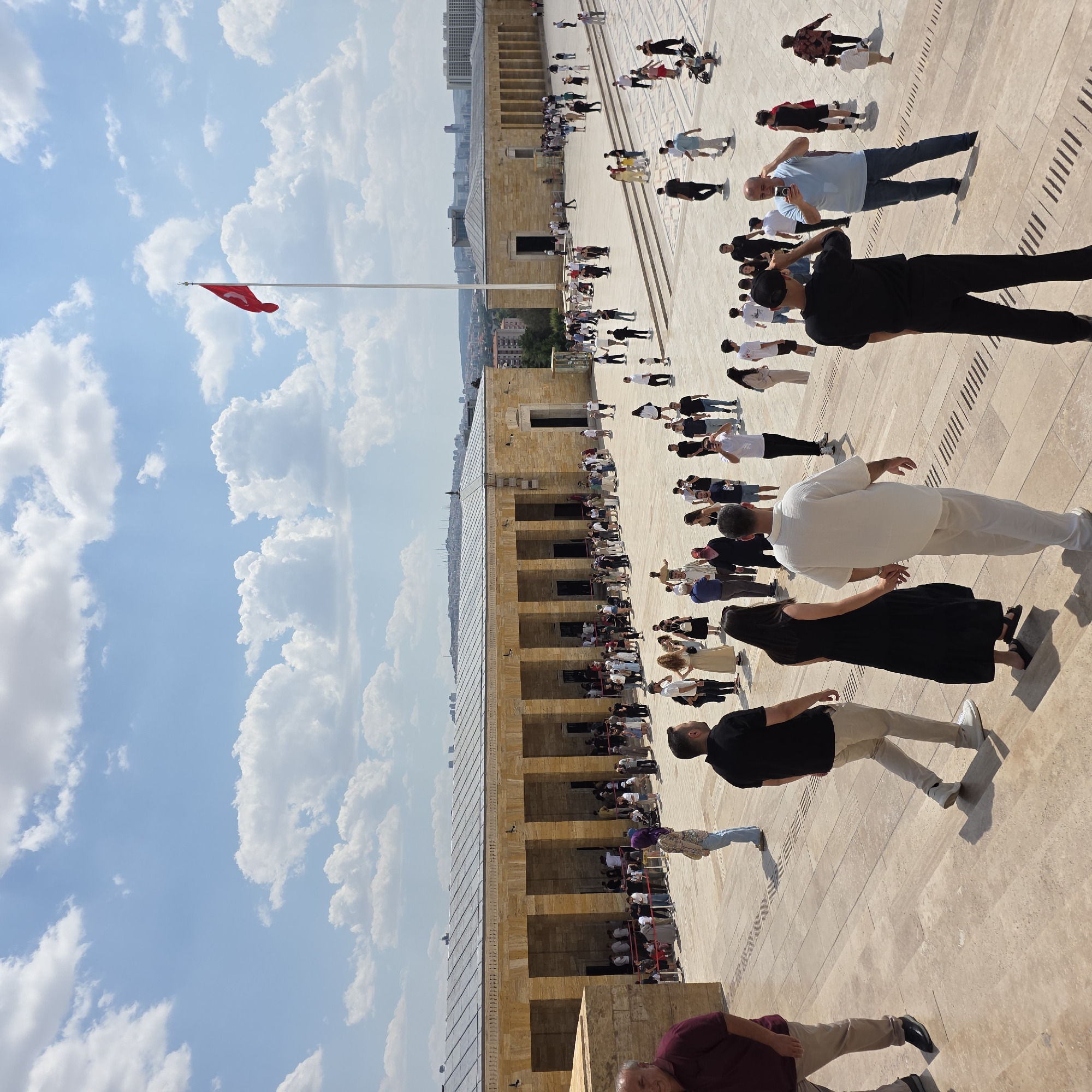 This is a wide-angle, eye-level shot of a large, open plaza in front of a stone building with a colonnade. The building appears to be a monument or historical site, possibly Anıtkabir, the mausoleum of Mustafa Kemal Atatürk, in Ankara, Turkey, given the architectural style and the presence of a Turkish flag on a tall flagpole in the background. The sky is blue with scattered white clouds.
There are many people in the plaza, most of whom are dressed in casual summer clothing. Many are standing in lines, suggesting they are queuing to enter the building or to view an exhibit. Some individuals are walking, and others are standing around.
In the foreground, on a stone staircase, a man in a maroon shirt and tan pants is walking towards the left. To his right and slightly behind him, a couple is walking down the stairs, holding hands. The woman is wearing a long black dress, and the man is in a white short-sleeved shirt and white pants. Further to the right, a man in a black polo shirt and tan pants is walking away from the camera.
In the midground, more people are visible. Some are in queues along the front of the building. A man in a light blue t-shirt and blue jeans is standing in the plaza, holding a smartphone and appearing to take a picture or video. Another man in a black t-shirt and black pants is standing in front of him, facing away from the camera.
The architecture of the building is characterized by its light-colored stone facade, rows of tall rectangular columns, and a flat roof with visible solar panels or lighting fixtures. The plaza is paved with light-colored stone slabs. In the far background, the cityscape of Ankara can be seen with buildings and structures on rolling hills under the sky.