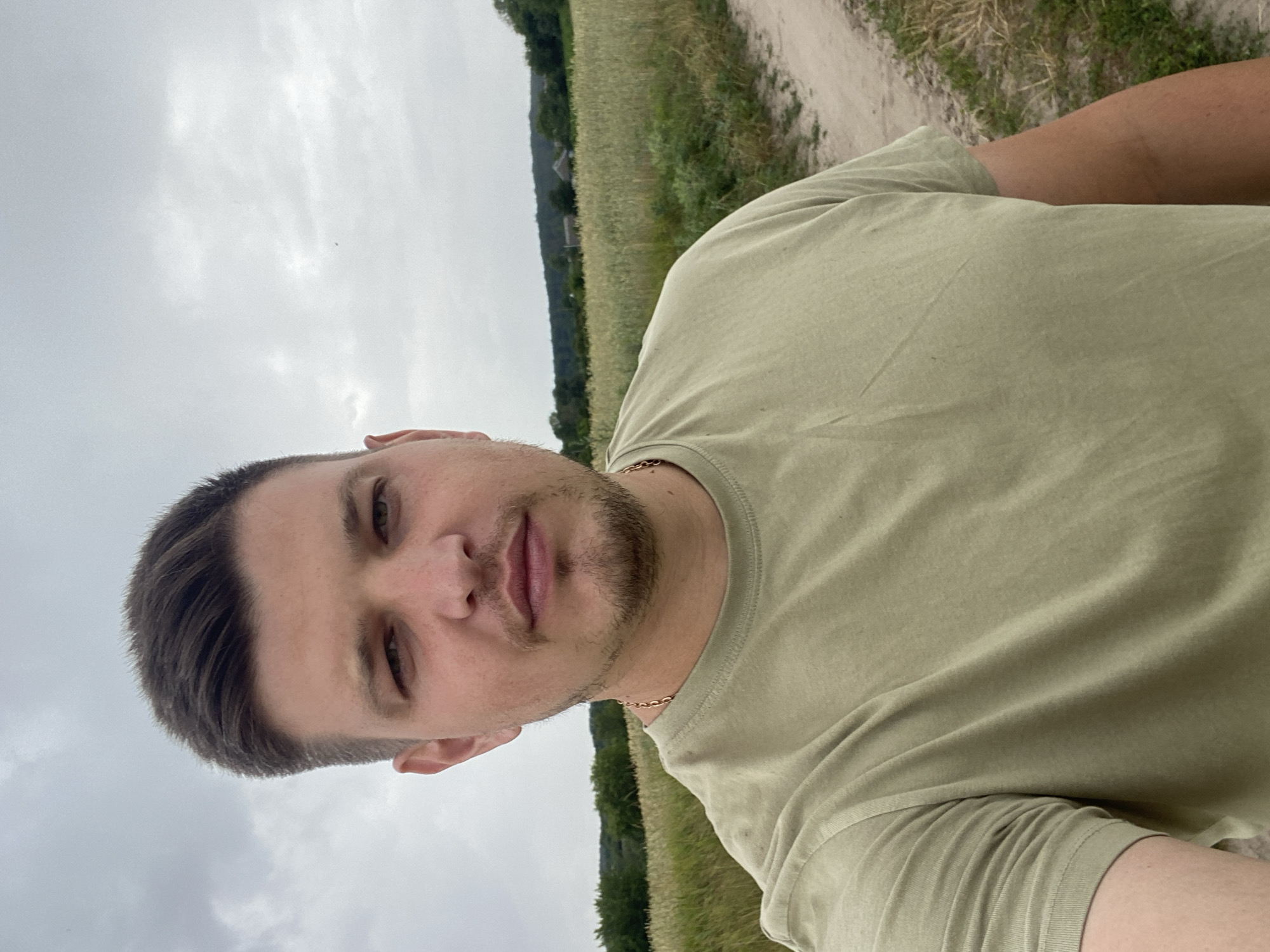 A man is standing in a field with a dirt path next to him. He is wearing a light green t-shirt and a gold chain. He has short brown hair and stubble on his face. The field appears to be wheat or barley. There are trees and some buildings in the background. The sky is overcast. The location is Yablunivka, Ukraine.