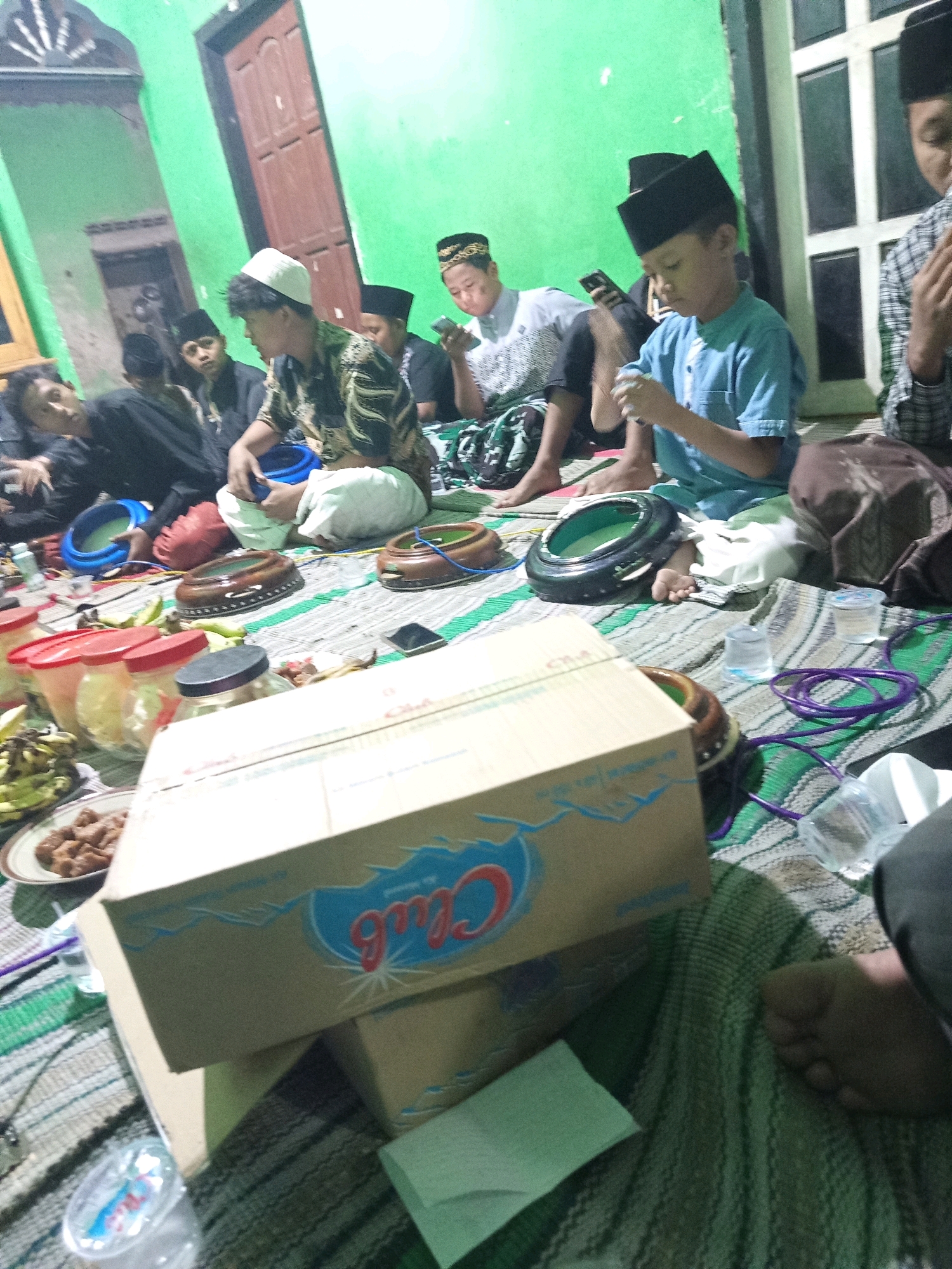 The image depicts a gathering of young boys and men indoors, possibly in Mrican, Indonesia, based on the provided context. The scene is dimly lit, with bright green walls forming the backdrop. Several individuals are seated on the floor, which is covered with mats.
In the foreground, a large cardboard box with the "Club" logo is prominently displayed. Next to it are various containers, some with red lids, suggesting food or drinks. There are also two wooden percussion instruments, resembling tambourines, on the mat. A purple coiled cable is visible near these instruments.
The people in the image are mostly young, wearing caps and traditional Islamic clothing. Some are looking at their phones, while others appear to be engaged in conversation or observing their surroundings. One individual in the center is wearing a blue shirt and appears to be holding something in his hands. Another person on the left is lying down, seemingly relaxed.
The overall atmosphere suggests a casual gathering or a small event. The presence of food containers and traditional musical instruments hints at a cultural or religious context. The room has a wooden door and a window with multiple panes on the right side. The lighting appears to be artificial, casting shadows across the scene.