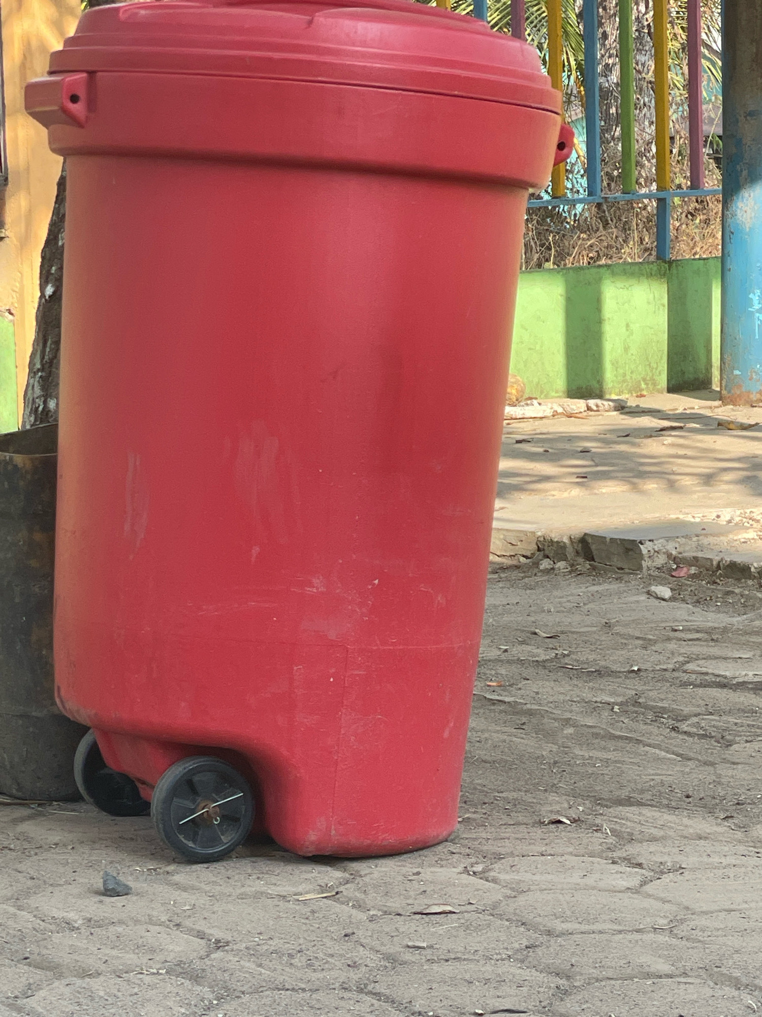 A bright red, wheeled trash bin stands on a paved surface in what appears to be an outdoor public area. The bin is large and cylindrical, with a domed lid. It has two black wheels on the bottom, one of which is clearly visible. The ground is covered in interlocking paving stones, with some loose gravel and dry leaves scattered about. In the background, there is a section of a green wall, a brightly colored fence with vertical poles of yellow, green, pink, and blue, and some foliage. The lighting suggests it is daytime, with shadows cast on the ground. To the left of the red bin, a portion of another dark-colored bin is visible. The overall scene is one of an urban or park-like setting, likely in the city of Bawahan Selan, Indonesia, based on the contextual information.