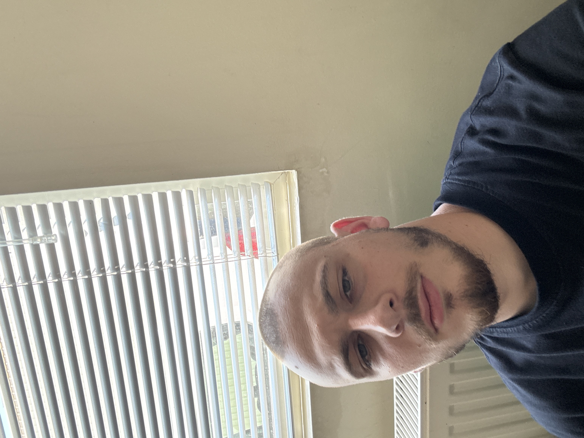 The image is a selfie of a young man with a shaved head and a goatee, wearing a black t-shirt. He is indoors, in front of a window with horizontal blinds. To the left of the man, there is a white radiator. The wall behind him is a neutral, light color. There is no indication of the city of Stara Zagora, Bulgaria.