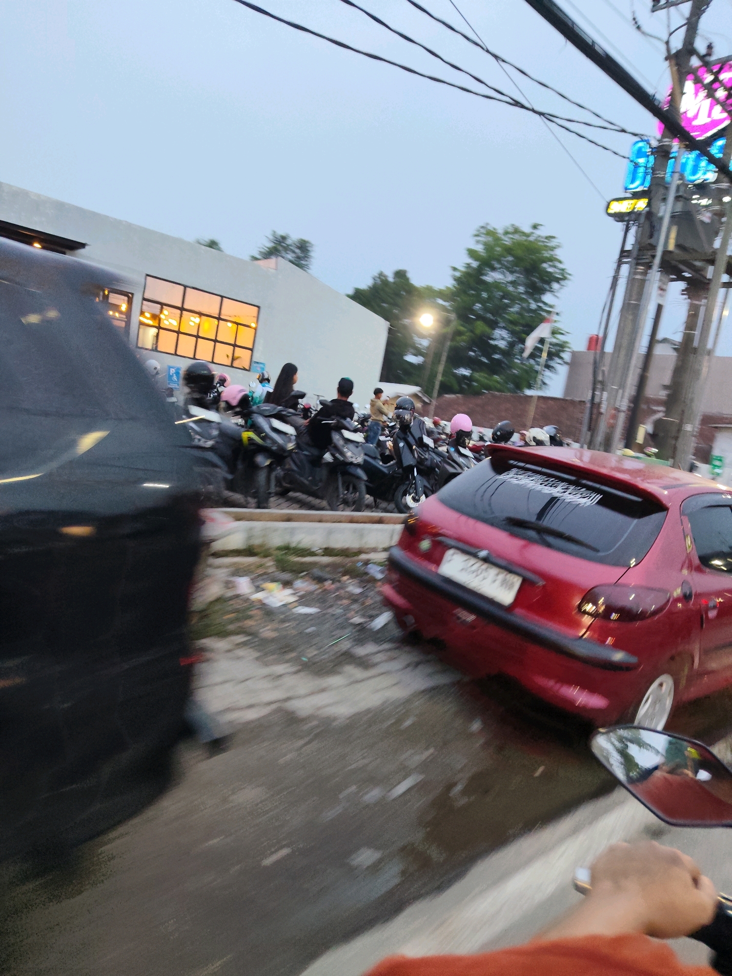 In Kaduagung Timur, Indonesia, a street scene unfolds with a blend of parked motorcycles and a red hatchback car. The motorcycles, predominantly dark-colored with some featuring bright pink helmets, are clustered together, suggesting a busy gathering or parking area. A red Peugeot 206 hatchback is parked on the right side of the image, its rear facing the viewer. A license plate is visible on the car, though the numbers are blurred. On the left, the back of a black car is prominently featured, its surface reflecting some of the ambient light. In the background, a modern building with large, amber-lit windows stands out against a muted sky. Overhead, power lines crisscross the frame, and a neon sign with blue and pink lettering is partially visible atop a pole. The ground appears wet and shows scattered debris, possibly indicating recent rain or a cleaning effort. The overall impression is one of a lively, possibly commercial, area with vehicles and a building contributing to the urban environment.