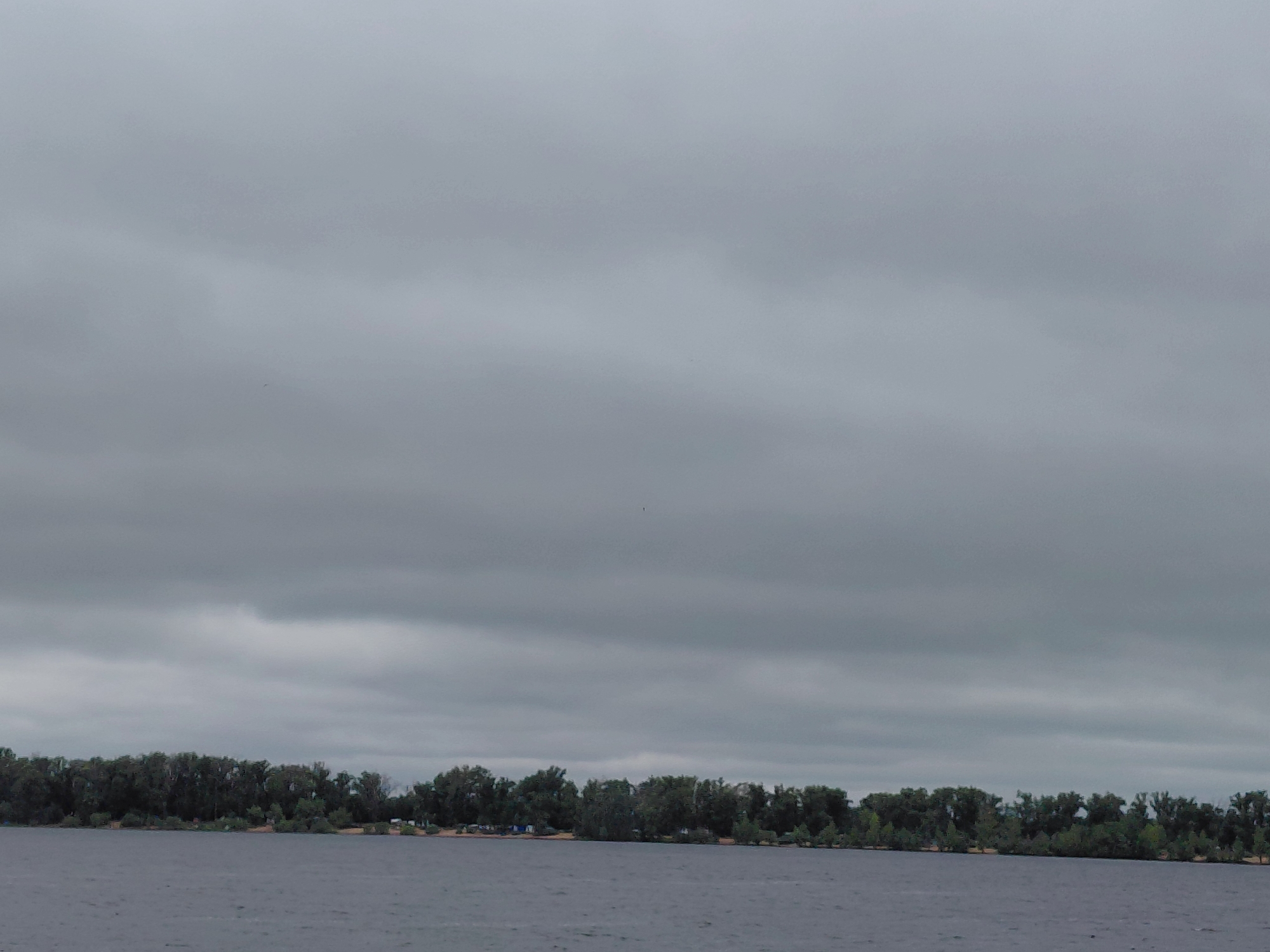 An overcast sky hangs low over a wide body of calm water, likely a river or lake. In the distance, a dense line of dark green trees forms a natural border along the far bank. Beneath the trees, a strip of lighter-colored sand is visible, suggesting a shoreline. A few scattered tents or small structures can be faintly discerned amongst the trees, indicating a potential camping area or settlement. The overall mood is somber and quiet, with the muted colors of the sky and water contributing to a sense of stillness. The location is identified as Novokuybyshevsk, Russia.