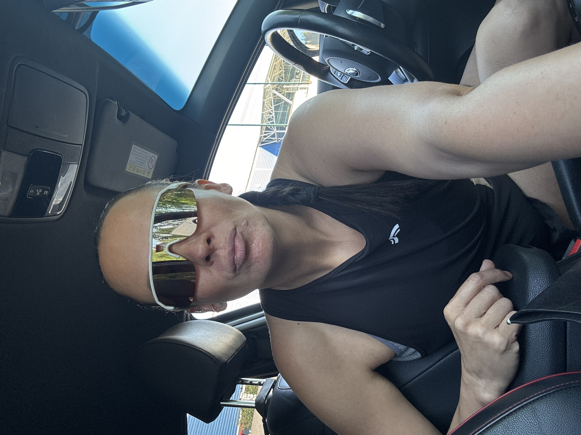 A woman in a black tank top and white-rimmed sunglasses takes a selfie in a car. She is in the driver's seat, with a black leather steering wheel visible. The sunroof is open, revealing a blue sky and what appears to be scaffolding. Based on the reflection in the sunglasses and the surrounding environment, the location is Chaikovsky, Russia.