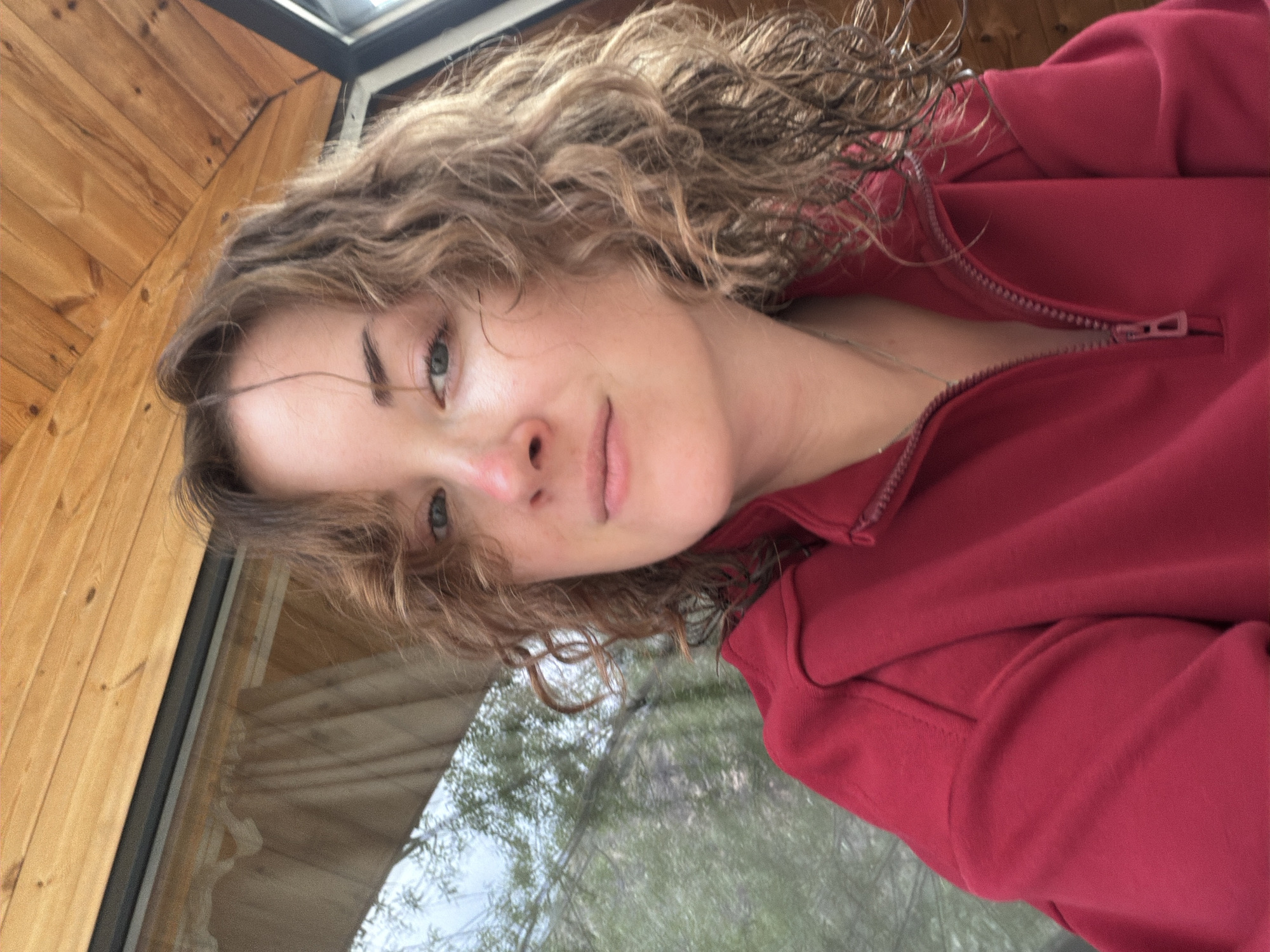 A close-up shot of a woman with curly hair wearing a red zip-up jacket, positioned indoors near a window displaying reflections of trees and foliage. The interior features wood-paneled walls. The location is Unknown, Unknown.