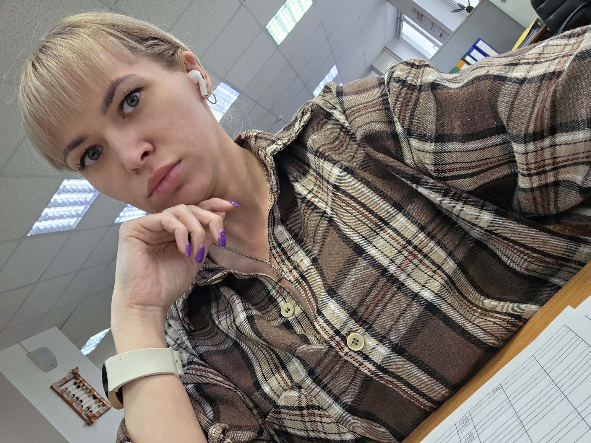 A young woman with short blonde hair and light-colored eyes is captured in a selfie, likely taken indoors in an office environment in Chaikovsky, Russia. She wears a brown, beige, and black plaid flannel shirt, with a silver chain necklace visible at her collarbone. Her left hand, adorned with sparkling purple nail polish, supports her chin, giving her a contemplative or serious expression as she looks directly into the camera.
She is accessorized with white wireless earbuds in her right ear and a cream-colored smartwatch on her left wrist. The background features a grid-patterned ceiling with fluorescent light panels, indicating an indoor setting. To her right, a wooden desk surface holds a stack of ruled papers, on which some numerical entries like "1380," "1300," "0101," and "100" are visible. In the further background, elements of an office are discernible, including a dark office chair, multi-colored folders or binders, and possibly a window. A small abacus is also visible on a white partition in the distance. The lighting suggests it is daytime.
