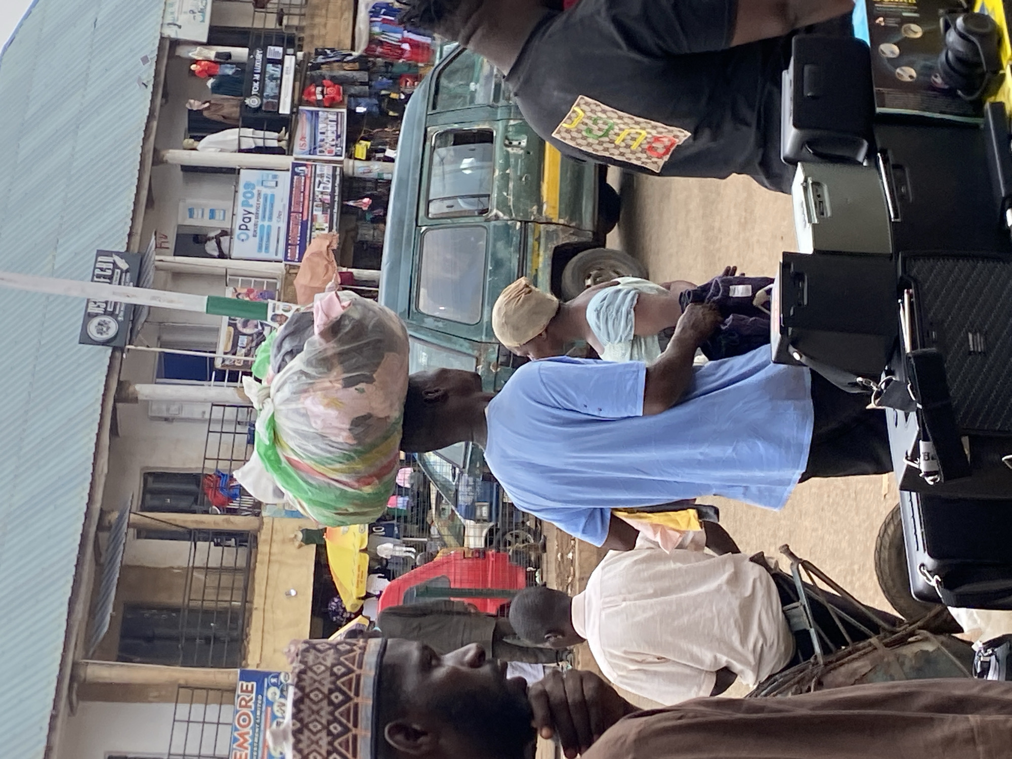 This image depicts a bustling daytime street scene in Bukuru, Nigeria, filled with people and commercial activity. The setting appears to be an outdoor market or a street lined with various shops and stalls.
In the immediate foreground, a man in a light blue t-shirt stands, expertly balancing a large, colorful bundle of what looks like textiles or goods on his head. To his right, another individual, possibly a child or a smaller adult, wearing a light blue garment and a light-colored cap, is bending over near a dark green van. Further to the right, a person in a black shirt, sporting a patch resembling "GUCCI" on their back, is partially visible, appearing to be attending to a stall laden with various black objects, possibly electronic devices or cases.
The background features several buildings with corrugated metal roofs, housing shops. Mannequins dressed in traditional attire are visible in one shop window. Several signs are prominent: "OPay POS BUKURU SERVICE POINT" indicates a financial service outlet, "OTOK M LUXURY" suggests a retail store, and another sign reads "EMORE." A green van, characteristic of local transport, is parked in the middle ground, adding to the urban landscape. The scene conveys a sense of vibrant daily commerce and movement in a lively community setting.