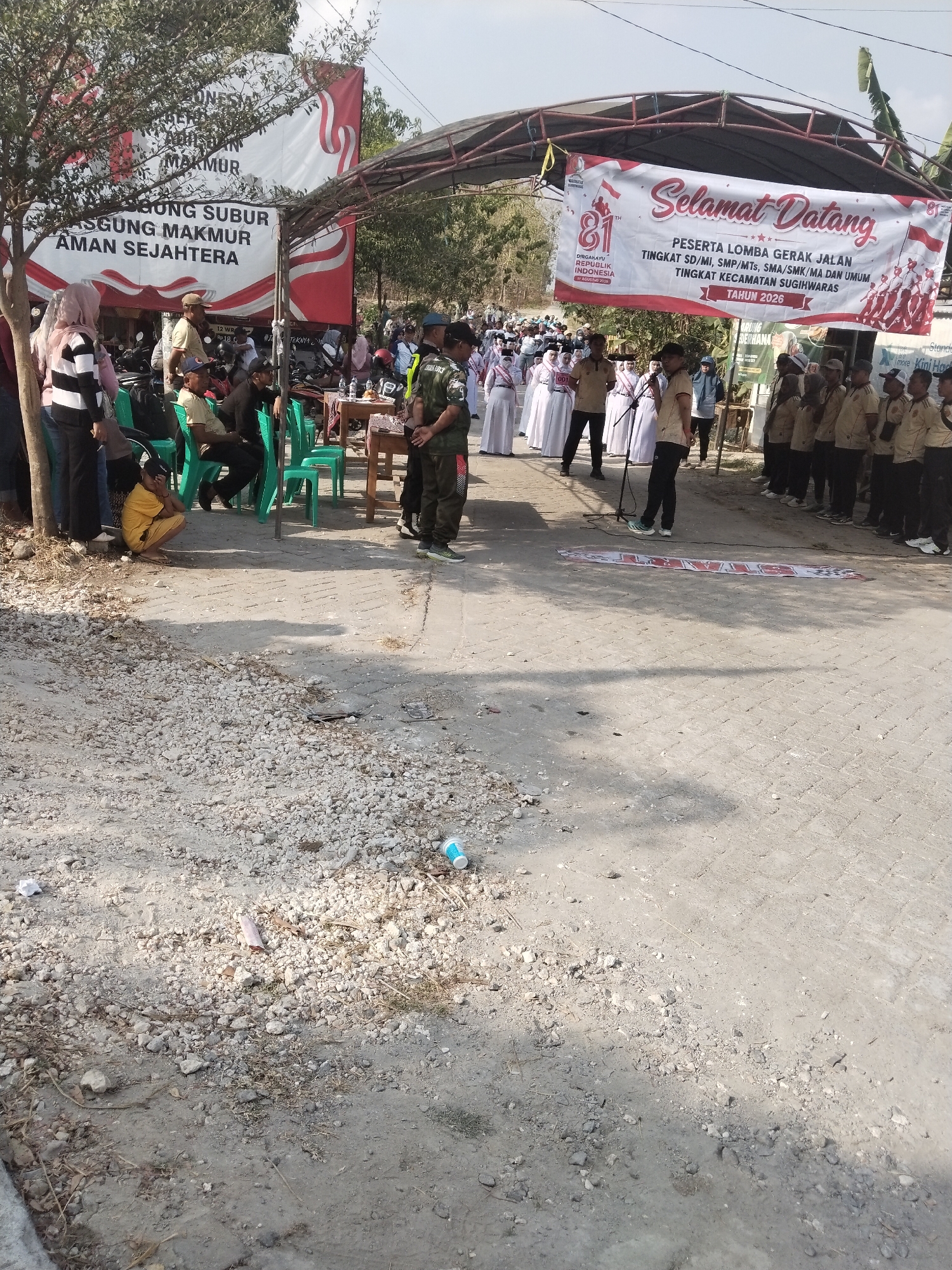 The image captures an outdoor event, likely a procession or competition, in the city of Alasgung, Indonesia. In the background, a large banner with red and white stripes reads "Selamat Datang" (Welcome) and advertises an event for "PESERTA LOMBA GERAK JALAN TINGKAT SD/MI, SMP/MTS, SMA/SMK/MA DAN UMUM TINGKAT KECAMATAN SUGIHWARAS TAHUN 2026." This translates to "Participants of the Road March Competition at the Elementary/MI, Junior High/MTs, High School/SMK/MA, and General Levels for the Sugihwaras Sub-district in the Year 2026." The banner is suspended from a makeshift structure.
In the mid-ground, a group of people in uniform, possibly students or participants in the event, stand in formation. A man in a military-style uniform stands to the left of this group, facing them. Another group of people in similar uniforms are visible to the far right, also seemingly participating in the event.
The foreground and surrounding area show a dirt and gravel path, indicative of an unpaved or developing area. Some trees are present, casting shadows on the ground. Scattered debris, including a plastic bottle and other small objects, are visible on the ground. A few people are seated on green plastic chairs to the left of the frame, observing the proceedings. A young person in a yellow outfit is crouching on the ground, partially obscured by a tree. The overall atmosphere suggests a community event taking place during daylight hours.