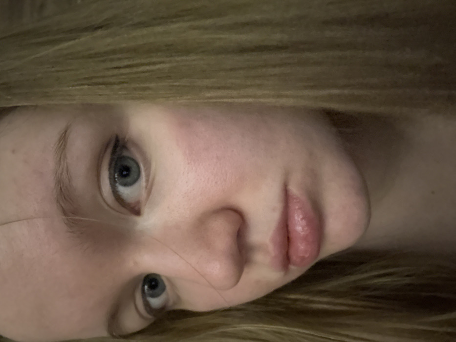 A close-up portrait of a woman with light skin and blonde hair framing her face. She gazes directly at the viewer with blue eyes. Her lips are full, and her nose is slightly upturned. The image has a soft, natural light, highlighting the texture of her skin. The composition is tilted, adding a slightly disorienting but intimate feel to the photograph.