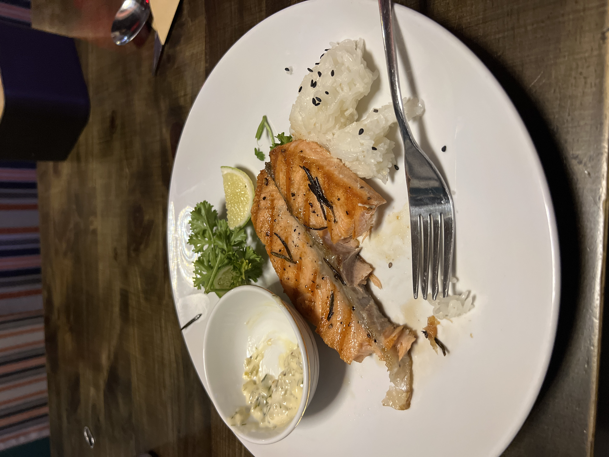 A close-up shot shows a partially eaten meal on a white plate, set on a wooden table. The main element is a piece of cooked salmon, seasoned with what appears to be herbs. Next to the salmon is a small white bowl containing a creamy sauce, possibly tartar sauce. A wedge of lime and sprigs of parsley garnish the plate. Sticky rice with black sesame seeds is also present. A silver fork rests on the plate, partially obscuring the rice. The meal is situated indoors. Location: Unknown, Unknown.