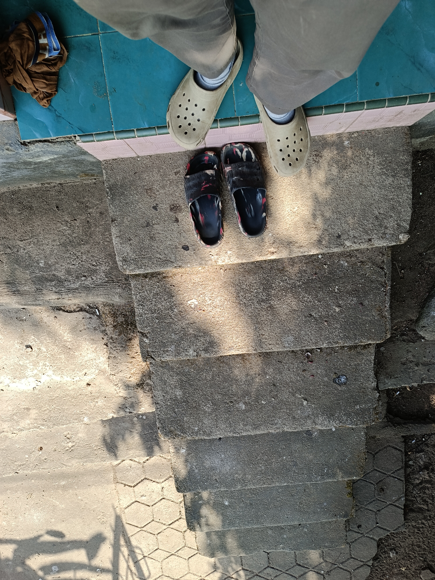 The image depicts a pair of feet wearing beige Crocs and a pair of black and red patterned slide sandals, standing on a set of concrete steps. A brown, folded garment lies on a blue tiled surface to the left of the Crocs. The scene is outdoors, with sunlight casting shadows on the steps. The environment appears to be an exterior walkway or entrance, possibly in a residential or public area within Mansalean, Indonesia. The concrete steps are worn and show signs of age. The ground surrounding the steps consists of dirt and some hexagonal paving stones.