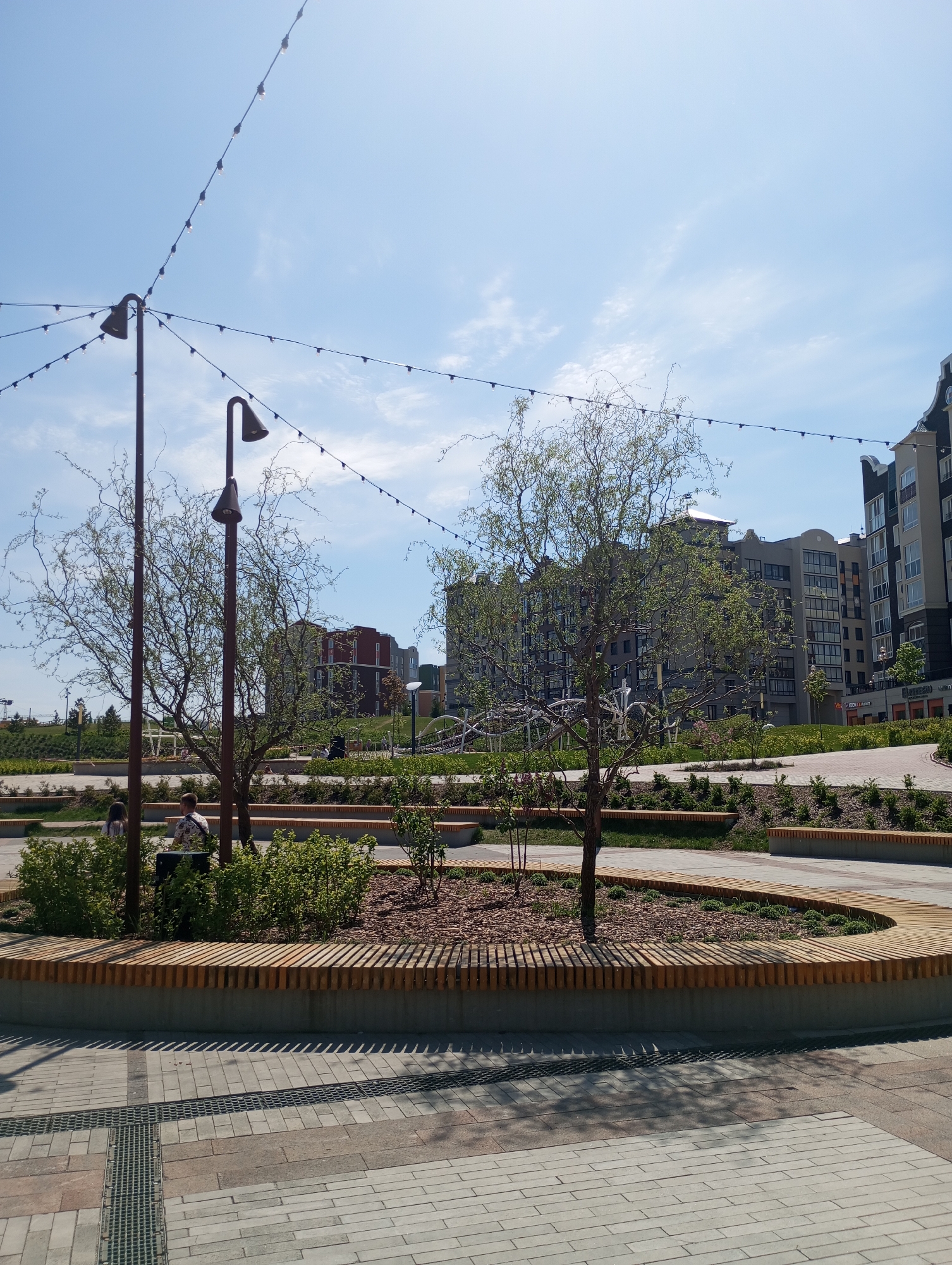 Captured on a sunny day in Izhevsk, Russia, the image shows a modern park area filled with landscaping and recreational features. In the foreground, a circular raised bed with wooden slats contains trees and small bushes. Two lampposts stand adjacent to the bed, and string lights are suspended overhead.
Two people are seated near the lampposts. The ground is paved with light-colored rectangular bricks.
In the background, several multistory apartment buildings are visible, completing the urban environment.