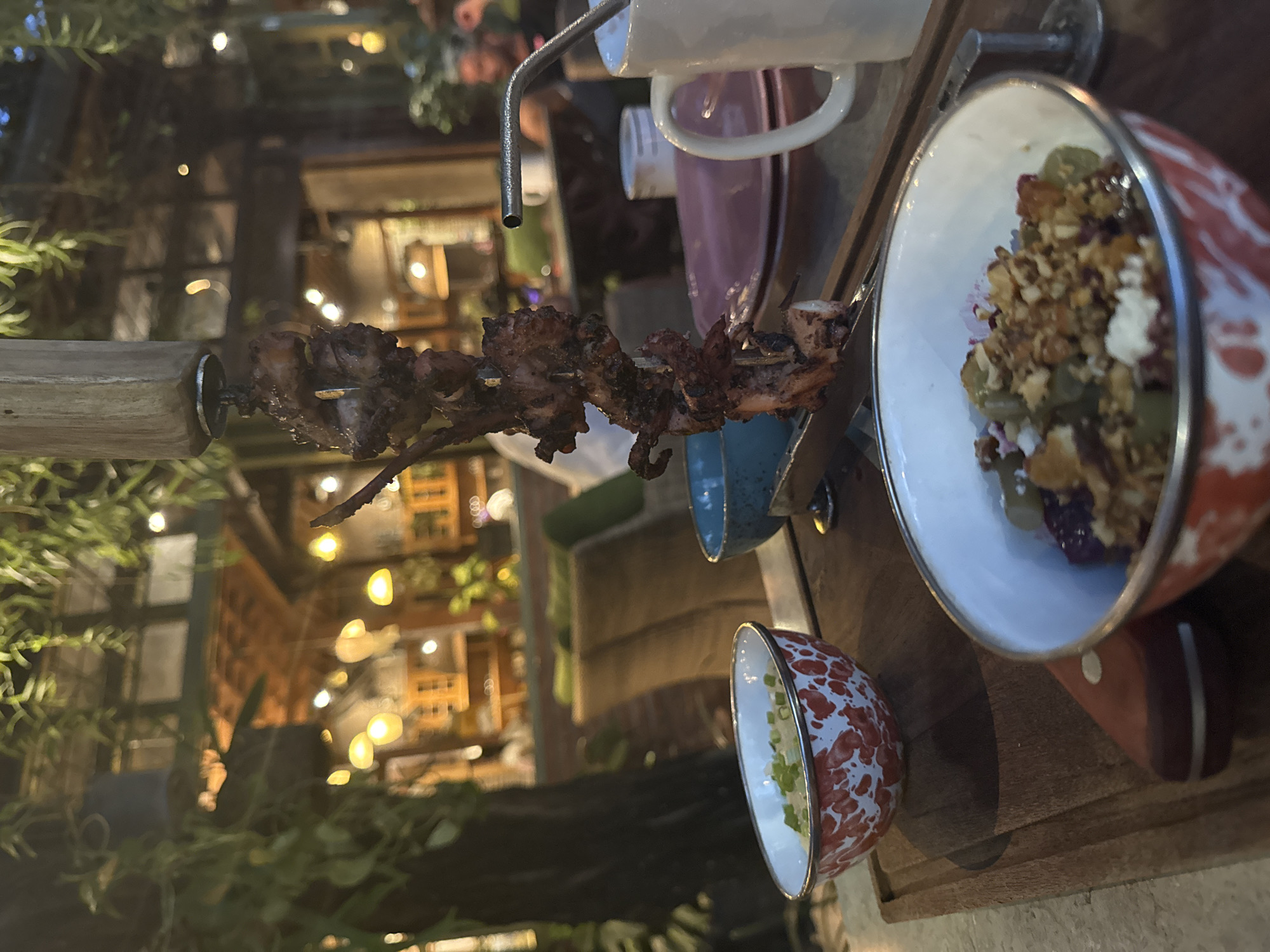 The image shows a table setting in what appears to be an outdoor restaurant or dining area in North Kuta, Indonesia. The table holds a skewer of grilled meat, several small bowls of food (one containing a salad with granola and other ingredients, and another with green onions), and various utensils and serving ware. The surrounding environment features foliage, decorative lighting, and glimpses of interior structures in the background.