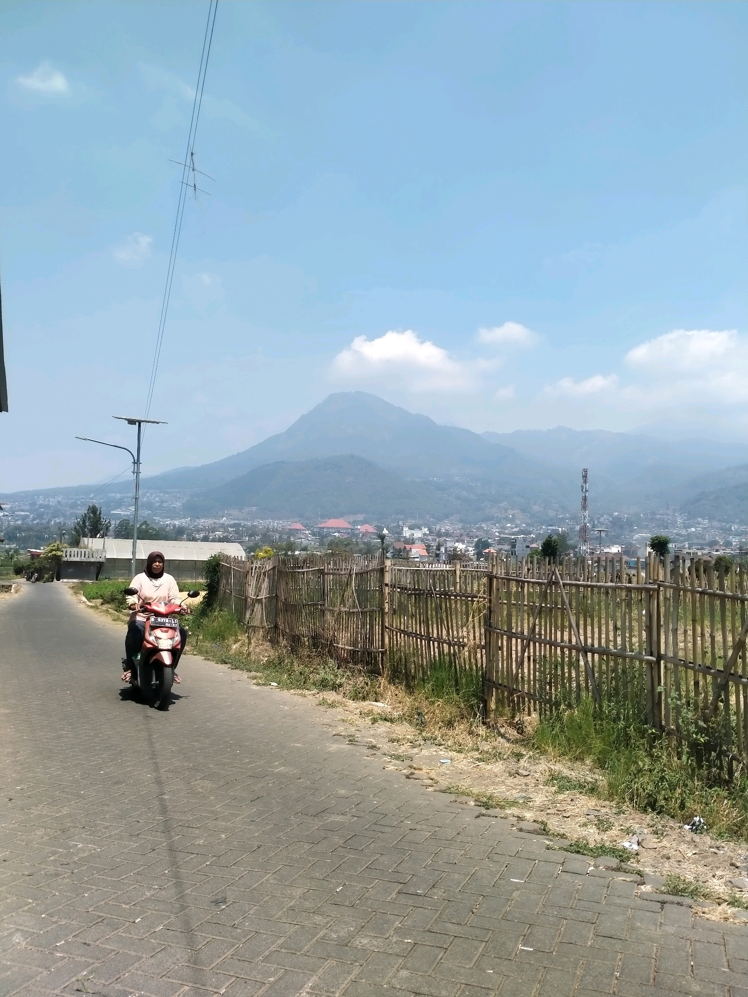 A woman wearing a headscarf and a light-colored shirt rides a red scooter on a paved road. The road has a cobblestone pattern and is lined with a bamboo fence on the right side. Beyond the fence, there is dry grass and some debris. In the background, a city is visible with buildings and a prominent cell tower. A large, hazy mountain range forms the backdrop to the city under a bright, clear sky with scattered white clouds. A street lamp with a solar panel is positioned on the left side of the road, near the woman. The overall impression is a daytime scene in a semi-urban or suburban area with a natural landscape in the distance.