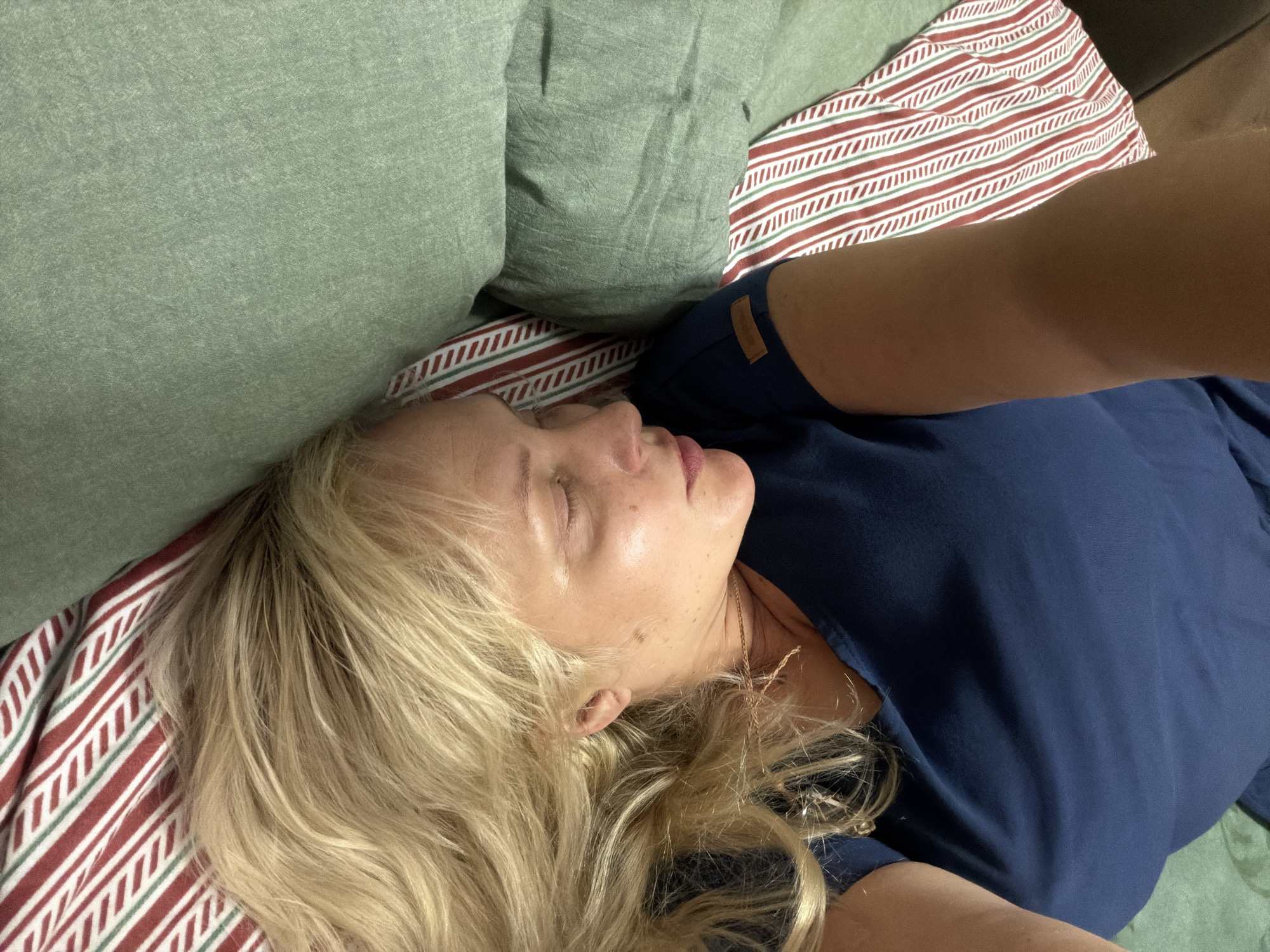 A woman with blonde hair is sleeping on a bed. She is lying on her right side, facing left. Her eyes are closed, and her mouth is slightly open. She is wearing a dark blue t-shirt. Her head is resting on a green pillow. The bed is covered with a striped blanket with red, white, and green stripes. The background is blurred and does not provide any contextual information about the location. There are no specific features that can identify the location as Kazan, Russia.