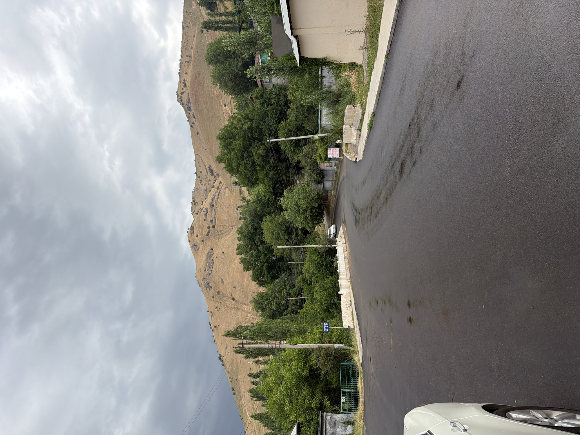 The image depicts a road leading towards a mountainous backdrop in Khumsan, Uzbekistan. The road is paved and appears wet, with reflections visible on its surface. On the left side of the road, there is a white car partially visible, along with a green metal fence and a concrete structure. Further down the road, there are utility poles with electrical wires. Trees and vegetation line the sides of the road, and in the distance, a hillside is visible, sparsely covered with dry grass and scattered bushes. The sky is overcast with heavy grey clouds. A sign with "HUMSON VILLA" written on it is visible near a utility pole. Buildings with simple architecture are present among the trees. The overall scene suggests a rural or semi-rural area on a cloudy day.