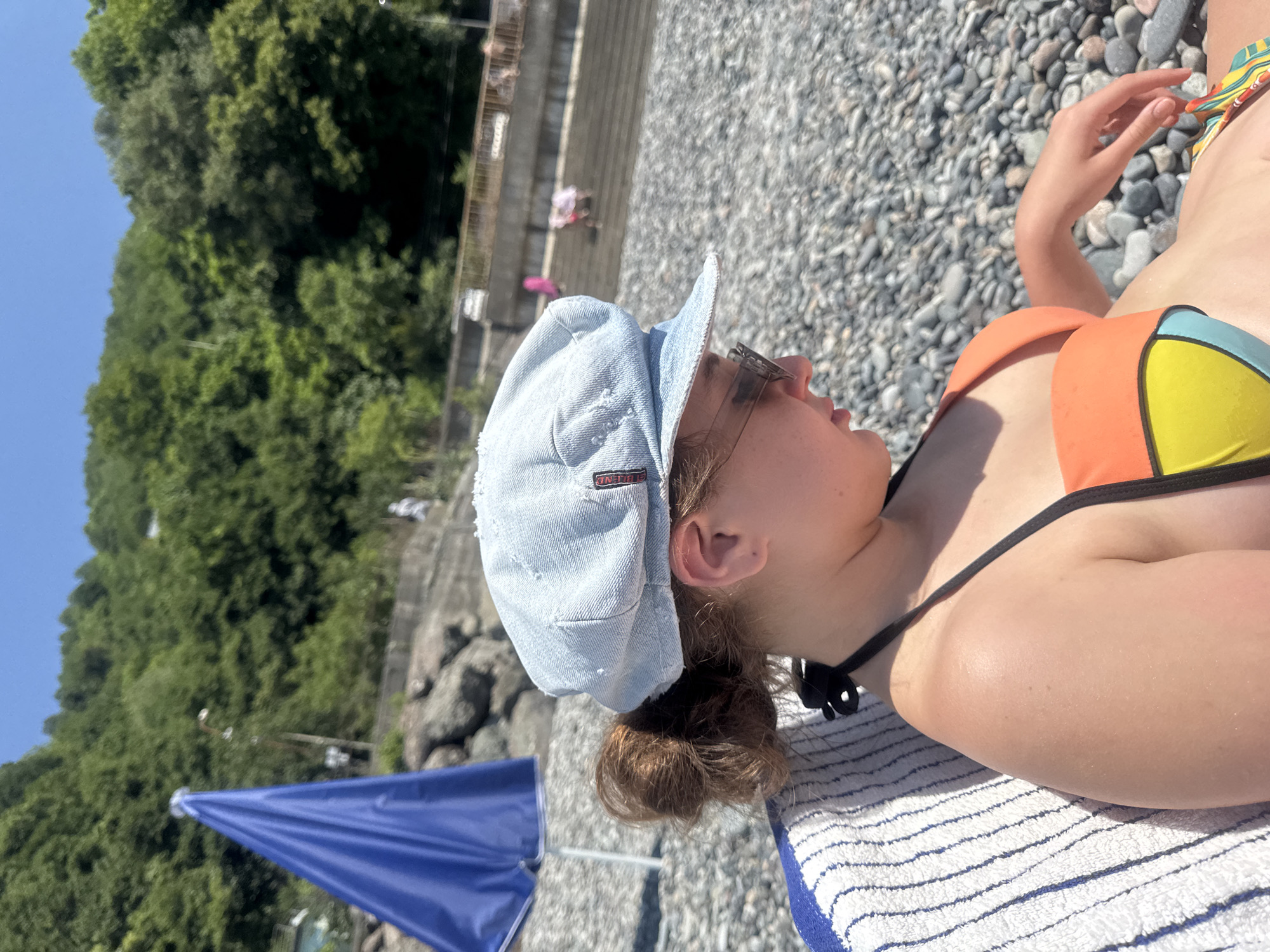 The media file captures a close-up of a young person lying on a towel on a rocky beach. The person is wearing a light blue denim cap and sunglasses, with their head tilted upwards and to the right, looking towards the sky. Their hair is pulled back into a bun. They are also wearing a colorful bikini with orange, yellow, and blue sections. A portion of a blue umbrella is visible in the lower left corner of the frame.
The background suggests a sunny day at the beach, with lush green trees on a hillside visible behind a retaining wall. The ground is covered in small, smooth pebbles. The lighting indicates it is likely daytime, possibly midday given the bright sunshine. The overall mood appears relaxed and summery.
A small tag on the denim cap is visible, although the text is difficult to read clearly. It appears to be a brand logo, possibly with some letters like "ON" and "BE".