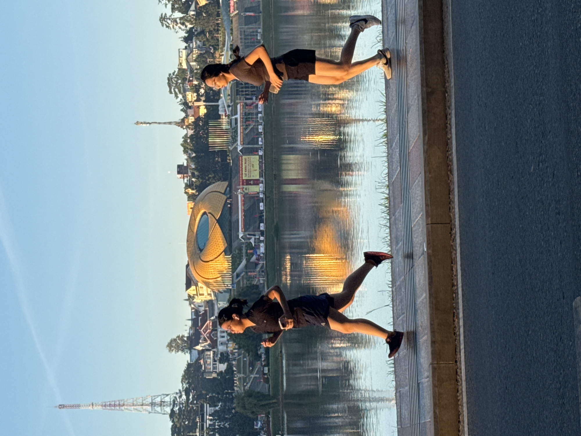 The image depicts two women running along the promenade of Xuan Huong Lake in Da Lat, Vietnam. The woman on the left, wearing glasses and a black t-shirt and shorts, is running with her left arm bent and her right arm holding a phone. The woman on the right, in a similar outfit, is running with her arms bent and holding a phone in her right hand. Both women are wearing athletic shoes.
The background features a picturesque cityscape of Da Lat, with several buildings, including a distinctive golden, dome-like structure, and a tall transmission tower. The lake itself is calm, reflecting the sky and the surrounding scenery. The promenade has a tiled surface with a grassy strip separating it from the lake. The scene is bathed in the warm light of either sunrise or sunset, casting long shadows and highlighting the golden hues of the buildings and the lake's surface. The road in the foreground is paved with asphalt.