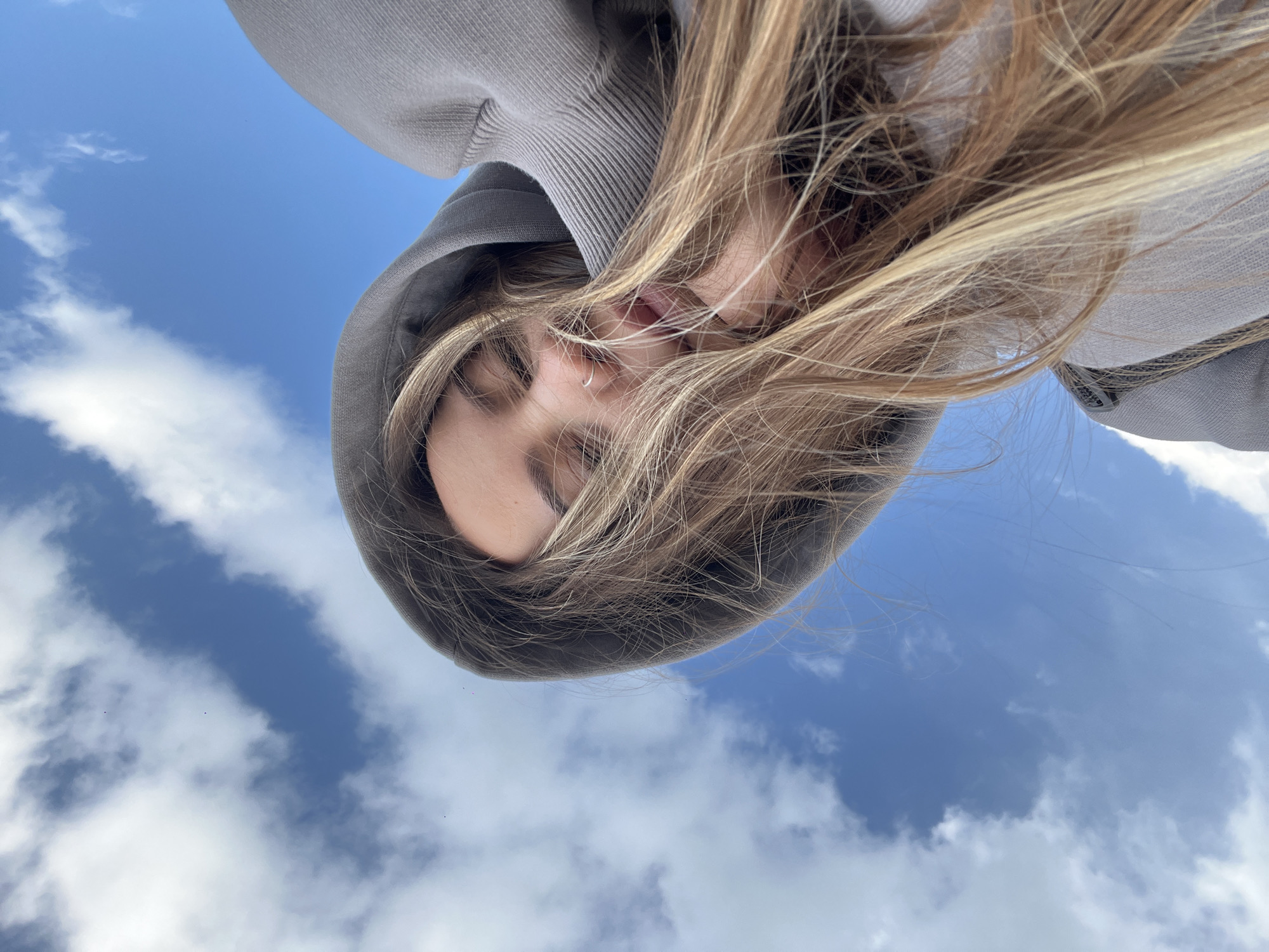 A female with long blonde hair is wearing a gray hooded sweatshirt. The view is from below looking up at her face. Her eyes are closed. The background is a blue sky with white clouds. There is no evidence of the location being in Novopokrovskaya, Russia.