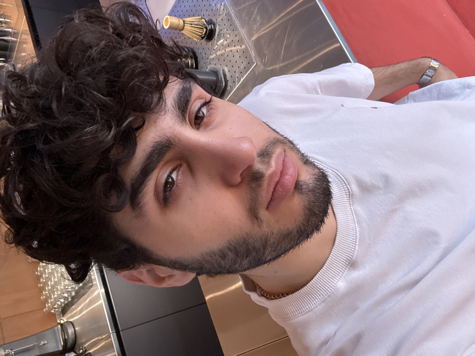 A young man with dark curly hair and a trimmed beard gazes at the camera. He wears a white sweatshirt and a gold chain. His expression is neutral, with full lips and dark eyes. The background features a metallic surface with a bamboo matcha whisk and a partially visible red wall. Lower left corner exhibits glasses and equipment on a black and metal countertop.