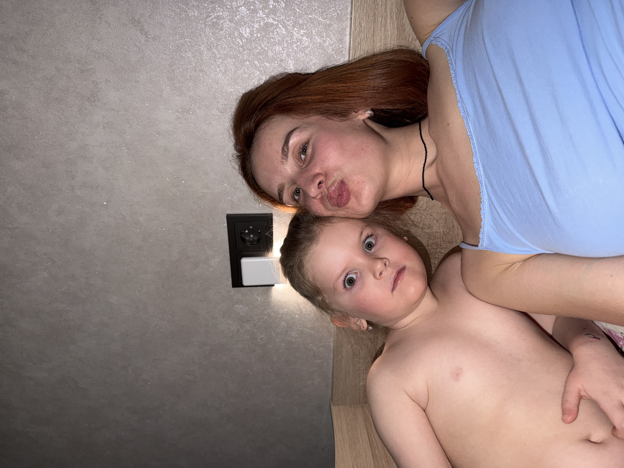 A photograph shows a young woman and a child posing for a selfie. The woman has red hair and is wearing a light blue top. She is making a kissing face toward the camera. The child is looking directly at the camera with wide eyes. The child is shirtless. They are indoors, possibly in a residential room, as indicated by the wall texture and the presence of an electrical outlet. The image appears to have been taken in Chaikovsky, Russia, as per the request.