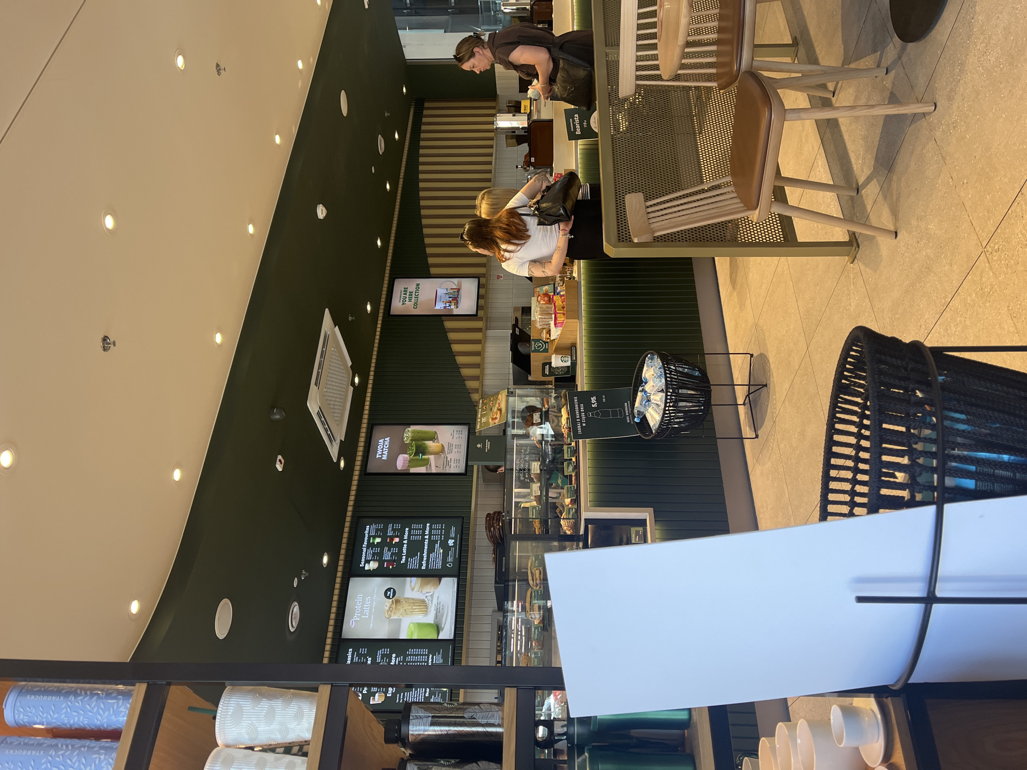 The image depicts the interior of a Starbucks coffee shop. The counter area is visible, with food displays behind glass cases. A menu board is displayed on the wall, featuring items like "Protein Lattes" and "Seasonal Favourites." Three women are present in the scene; two are standing at the counter, appearing to interact with staff, while another woman stands to the right, holding a bag and looking towards the counter. The environment is clean and modern, with green accent walls and wooden furniture. There are also tables and chairs in the foreground, suggesting a seating area for customers. Various Starbucks merchandise, such as cups and reusable bottles, are visible on shelves. A black basket filled with water bottles is situated near a white informational board. The lighting appears to be a mix of ambient ceiling lights and spot lighting on the counter and menu boards.
