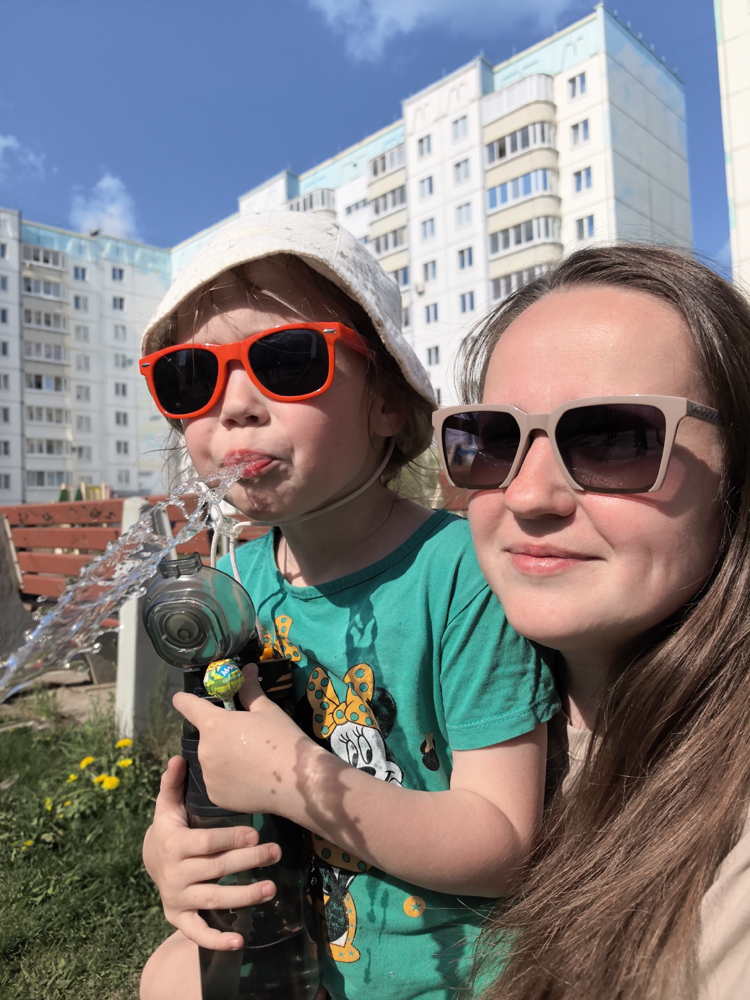 A medium shot shows a woman and a young girl standing outdoors with buildings in the background. The girl is wearing a white sun hat and red sunglasses and drinking water from a black and silver water gun that she holds. She wears a green t-shirt with a cartoon character on it. A lollipop is attached to the water gun. The woman is wearing beige sunglasses and has long brown hair. They are both facing forward toward the camera. Behind them is a grassy area with yellow flowers, a red wooden bench, and large, white apartment buildings with blue accents. The location is the city of Фролы, Russia. The sky is blue with some white clouds.