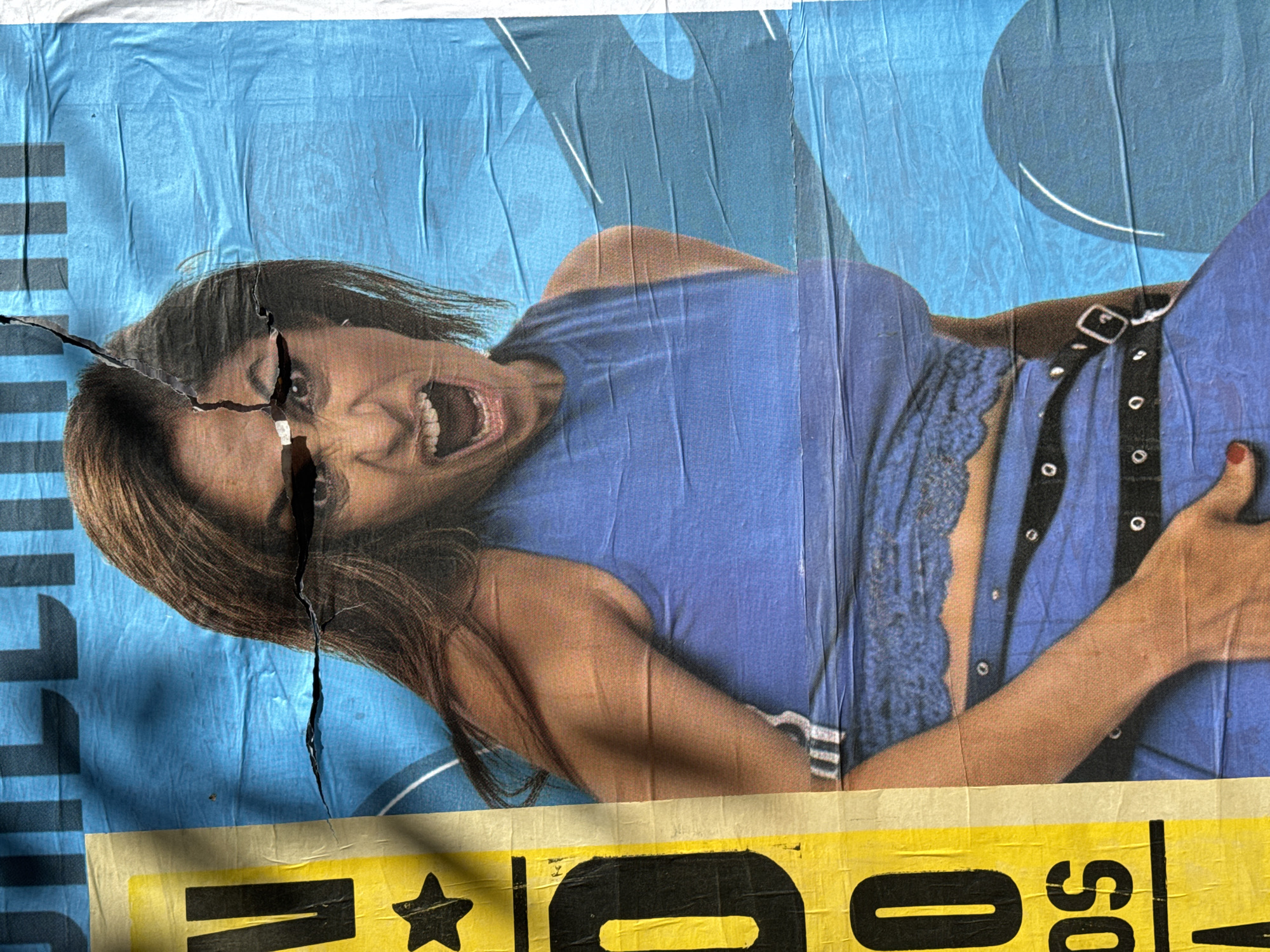A tattered, torn poster is adhered to a wall, likely in Buenos Aires, Argentina, showing a woman with brown hair, wearing a blue outfit and a black belt, lying down, shouting with her mouth open. The poster is blue and yellow, with some black text on the left side. The face of the woman is partially torn.