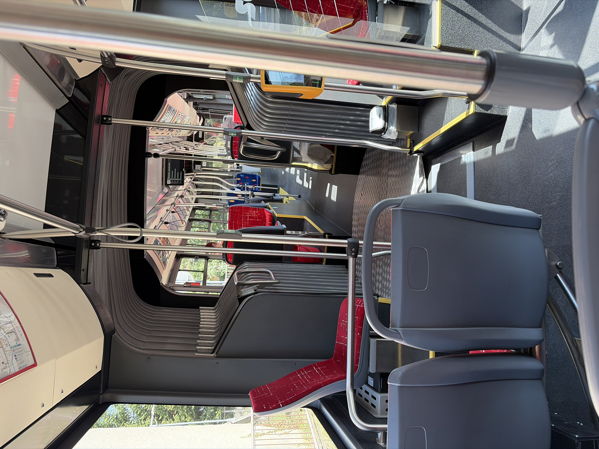 The image shows the interior of a bus, specifically the aisle and seating area, looking towards the back. The seats are a mix of red and blue, with some red seats featuring a checkered pattern. There are metal poles and handrails throughout the bus for passengers to hold onto. The floor is a dark, textured material. Natural light streams in from the windows on the left side of the bus, illuminating the interior and casting shadows. Outside the windows, trees and a fence are visible, suggesting a location in a greener area. A map is displayed above the windows on the left. The overall impression is of a modern public transport vehicle. The information on the digital display board visible in the distance indicates the location is within the city of Nitra, Slovakia.