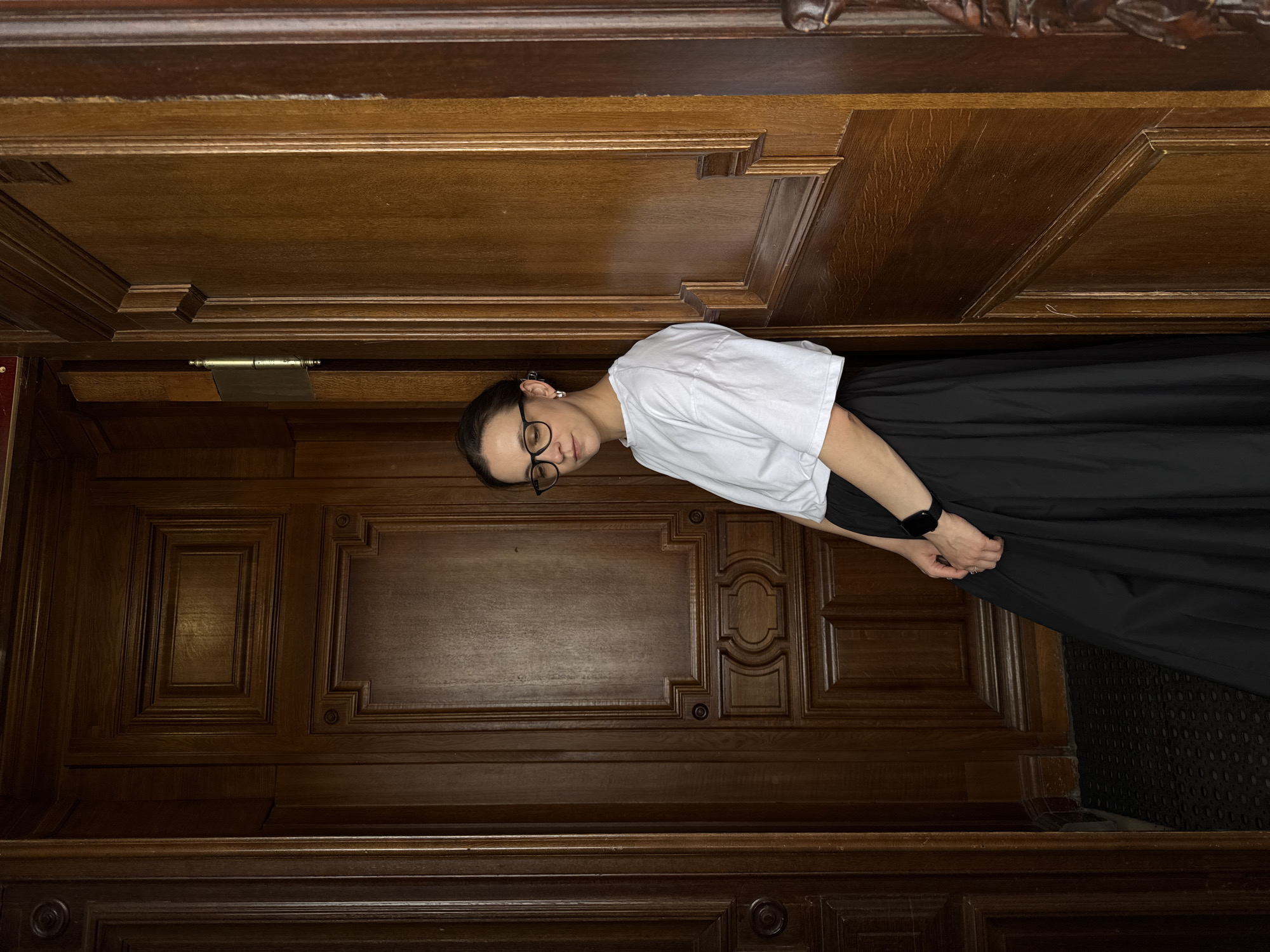 A person wearing glasses, a white t-shirt, and a long black skirt stands indoors, facing the camera. The background is a dark wood paneled wall with ornate carvings. The location is Nizhny Novgorod, Russia.