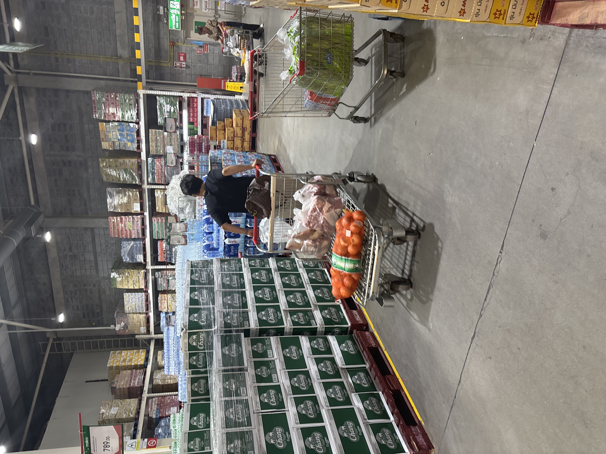The image shows an interior view of a Makro store, possibly in Baan Bo Phut, Thailand. It features rows of stocked shelves with a variety of beverages and products. In the foreground, there is a large display of cases of Chang beer. A person in a black shirt is pushing a shopping cart filled with groceries, including a bag of oranges and other items. Another shopping cart with produce is visible further away. The floor is concrete and there are price tags and promotional signs visible near the displays. The overall scene depicts a typical shopping environment inside a wholesale store.