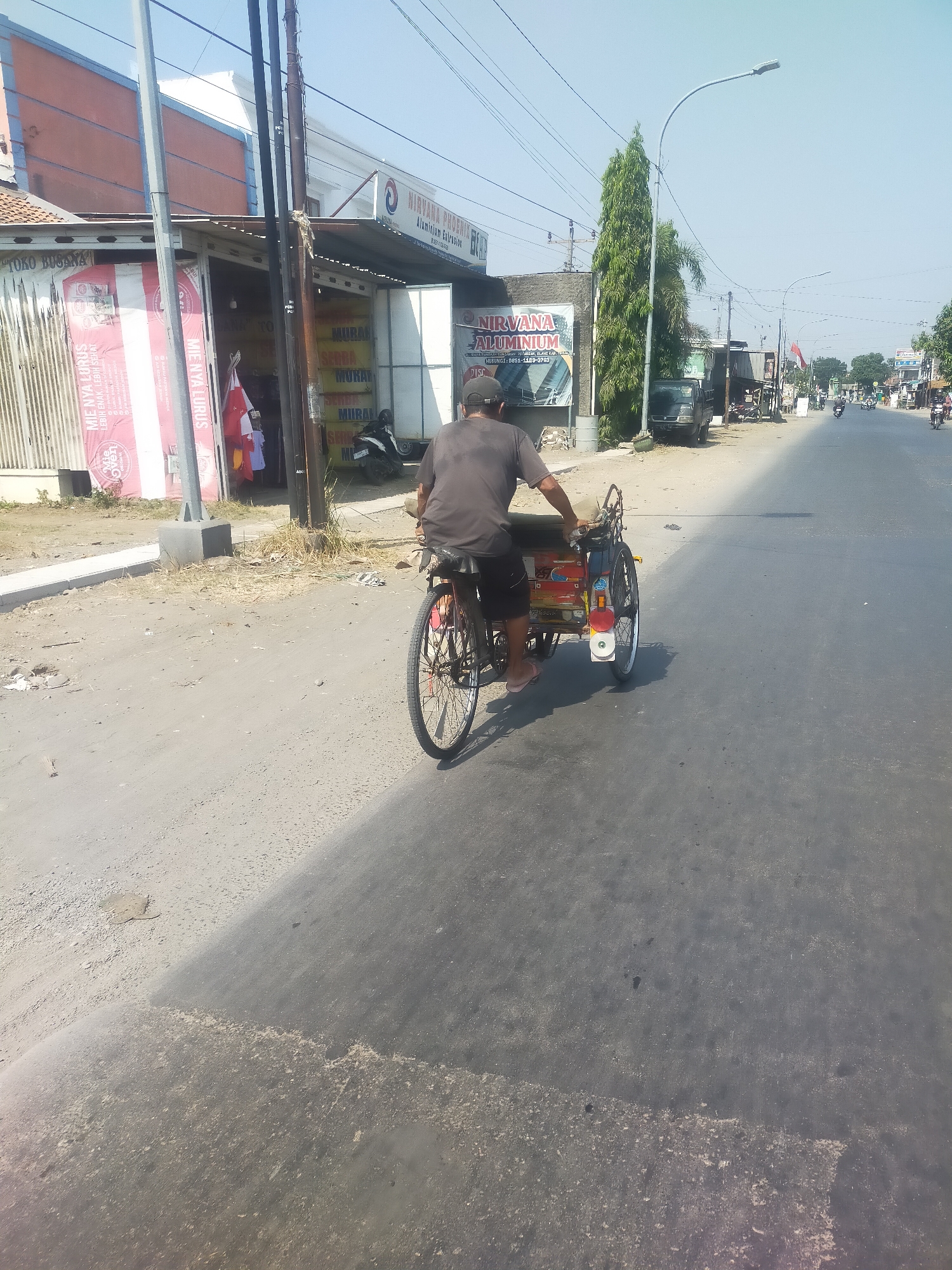 In Pekiringan, Indonesia, a man is seen riding a three-wheeled bicycle with a passenger carrier attached. The man is wearing a dark gray t-shirt and black shorts, with pink flip-flops. He is operating the bicycle on a paved road that is partially covered with dust and debris. The bicycle has two rear wheels and one front wheel. The passenger carrier is painted with various colors and designs, including red, yellow, and white. The scene appears to be on a street with shops and businesses lining one side. A tall, slender evergreen tree stands near the roadside, and a utility pole with wires is visible in the background. The sky is clear and blue, indicating a sunny day.