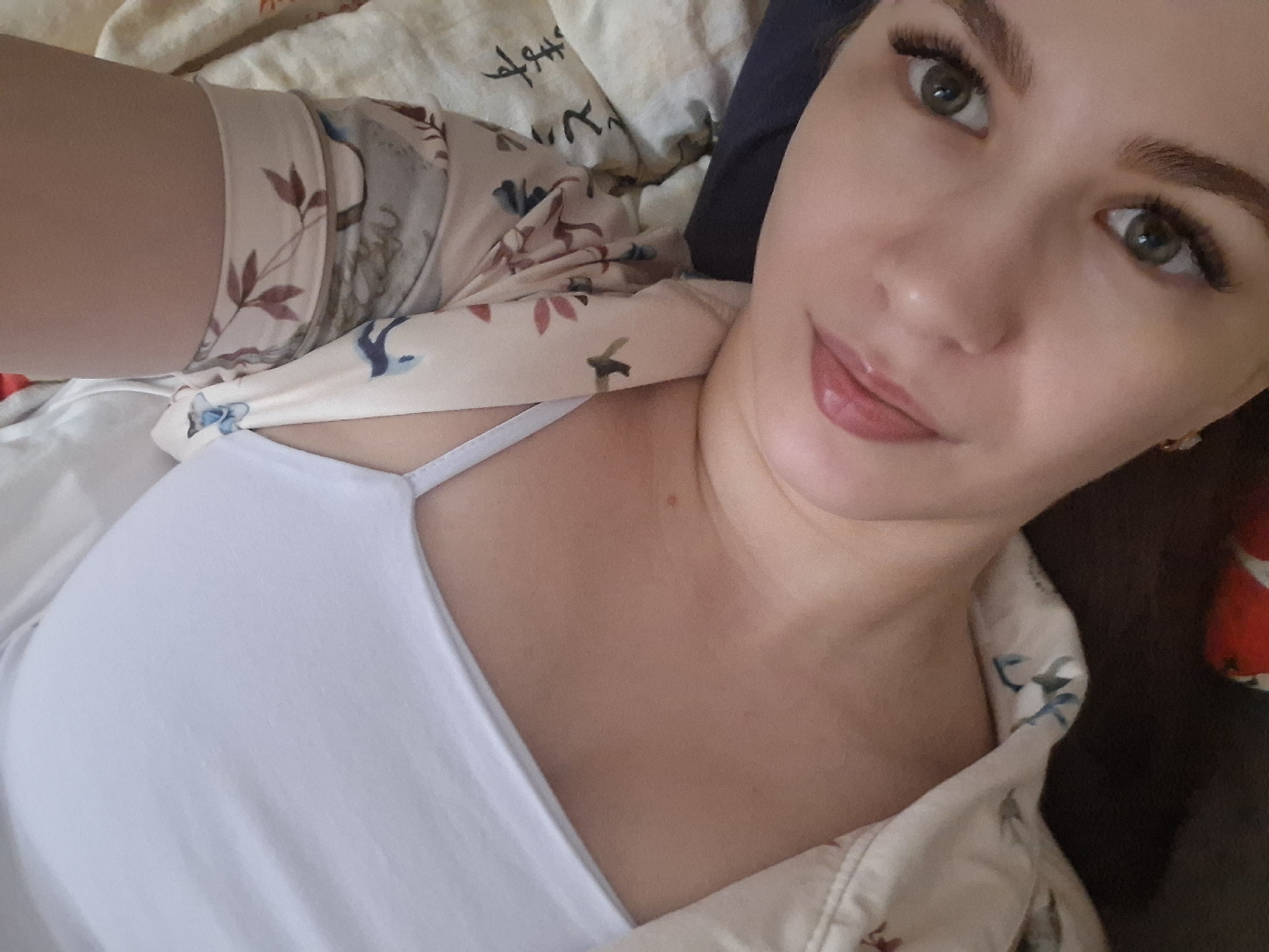 A person with light skin and green eyes is looking at the camera. They have long eyelashes and full lips. The person is wearing a white tank top and a patterned shirt. The background is a bed with a patterned sheet. The photograph appears to be taken in an interior location, with no other indications of the location in Chaikovsky, Russia.