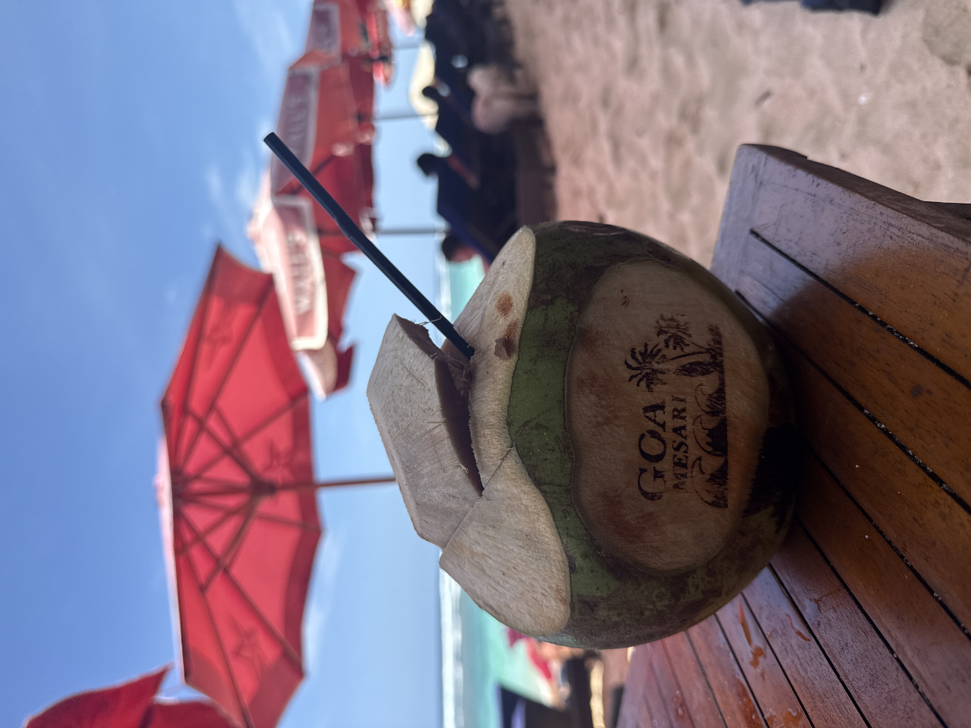 The image captures a coconut drink with a black straw resting on a wooden table, likely at a beachside establishment. The coconut is labeled "GCA MESARI". Red beach umbrellas are visible in the background against a blue sky, indicating a sunny day. There are people and sand in the blurry background suggesting a beach setting. The image is taken in Kuta Selatan, Indonesia.