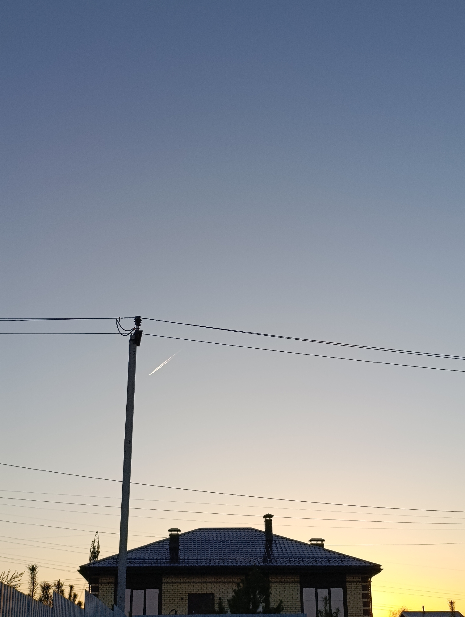The image shows a two-story house with a dark tiled roof, two chimneys, and visible windows at dusk. The house appears to be made of brick. A utility pole with attached power lines is in the foreground, slightly obscuring the view of the house. A contrail is visible in the sky behind the pole. The sky is a gradient of blue to yellow, indicating either sunrise or sunset. A white fence is visible on the left side of the frame. The location is presumed to be Chaikovsky, Russia.