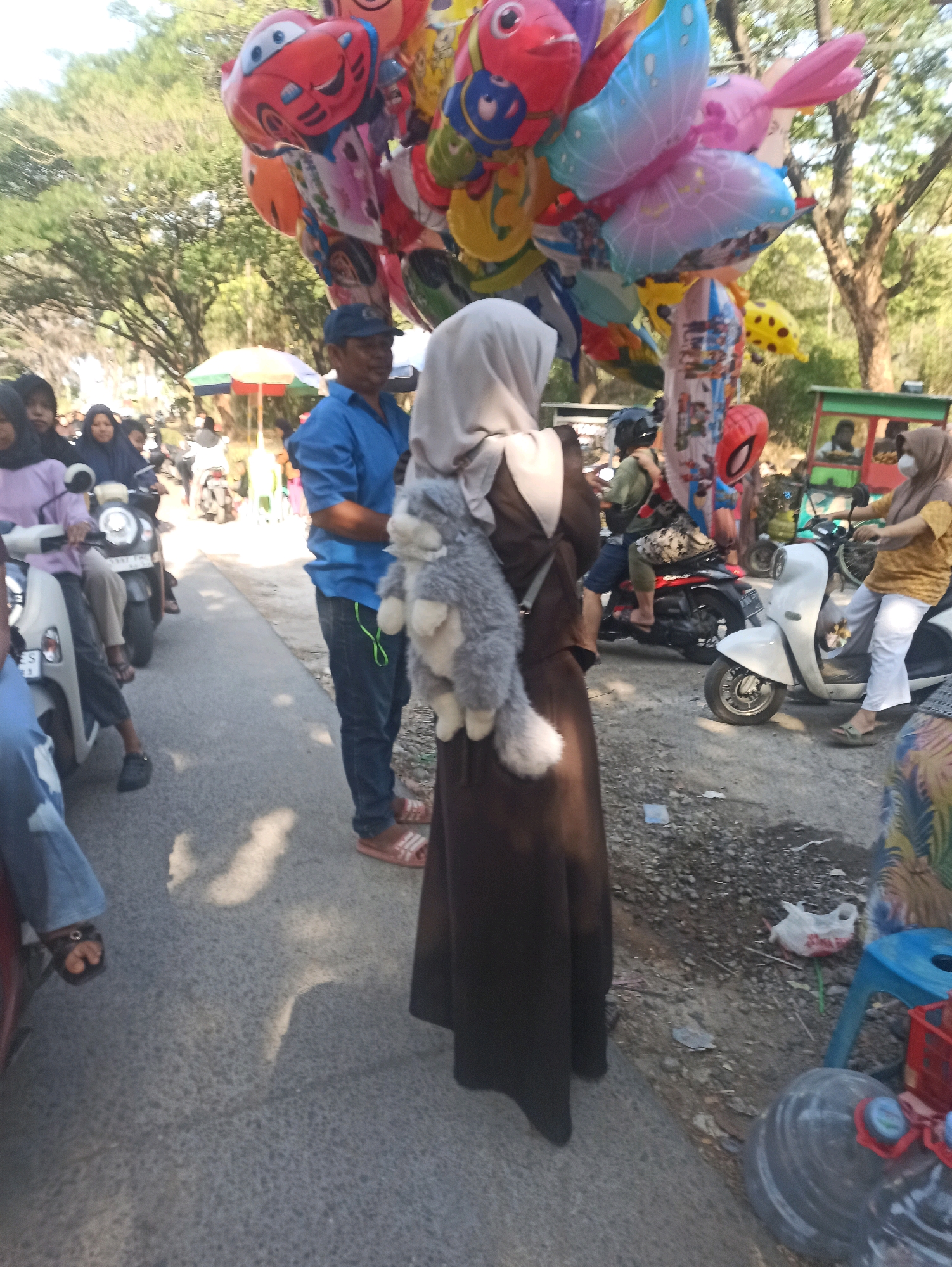 The image depicts a street scene in Telagamurni, Indonesia, likely during the daytime given the bright lighting and shadows. A variety of people are present, some on motorcycles and others standing. In the foreground, a person wearing a brown abaya and a light grey hijab is facing away from the camera, carrying a stuffed animal on their back. To their left, a man in a blue shirt and dark jeans is standing, looking towards the right.
Further back, several people are visible on scooters. One scooter has two individuals on it, one in a purple top and another in a dark hijab. Another scooter further down the road appears to have a driver and passenger wearing helmets. In the background, trees and some structures, possibly market stalls, are visible. A cluster of colorful balloons, including shapes resembling cars, butterflies, and cartoon characters, are held aloft, likely by a vendor. The road surface is asphalt, with some debris and shadows scattered across it. The overall impression is of a busy, casual gathering or marketplace.