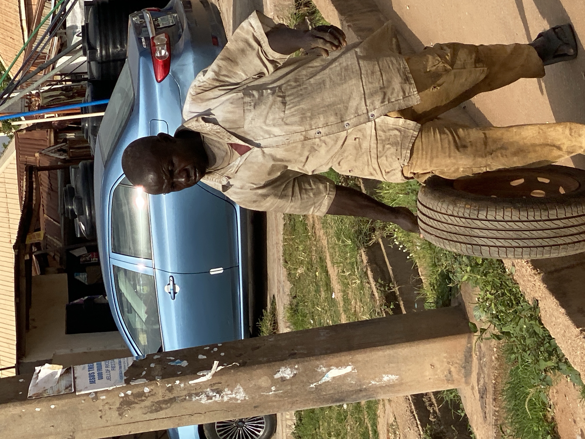 A man with dark skin walks forward carrying a tire. He is wearing a tan, short-sleeved, button-down shirt with a red undershirt visible at the collar. His pants are also tan and appear to be made of a sturdy material. He is wearing dark sandals on his feet. The man is in a dirt and grassy area with a concrete border. In the background, a blue sedan is parked, and further back, buildings with corrugated metal roofs are visible. Some black water tanks are also present. A wooden utility pole with posters attached stands to the left of the frame. The scene appears to be in a marketplace or a busy roadside area in Sabon Tasha, Nigeria.