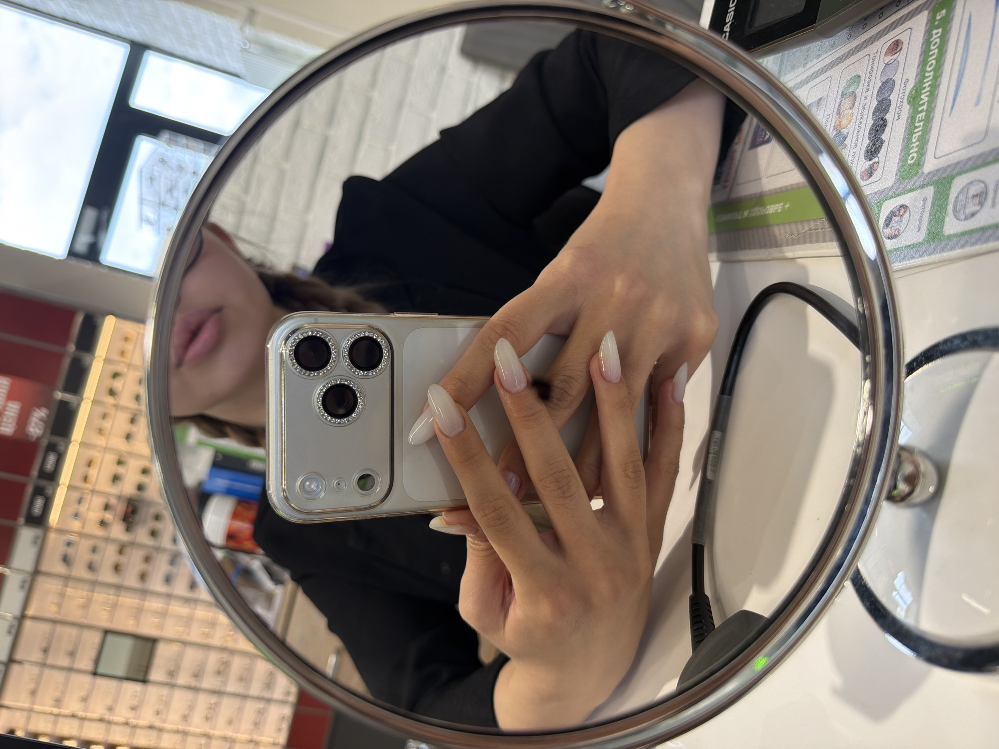 The image shows a person taking a selfie in a circular mirror. The person is holding a smartphone with a prominent camera array. The background, reflected in the mirror, shows shelves filled with eyewear, suggesting the location is an optician's shop or a store selling glasses. There's also a desk with papers and a CASIO device visible. The lighting suggests an indoor environment.