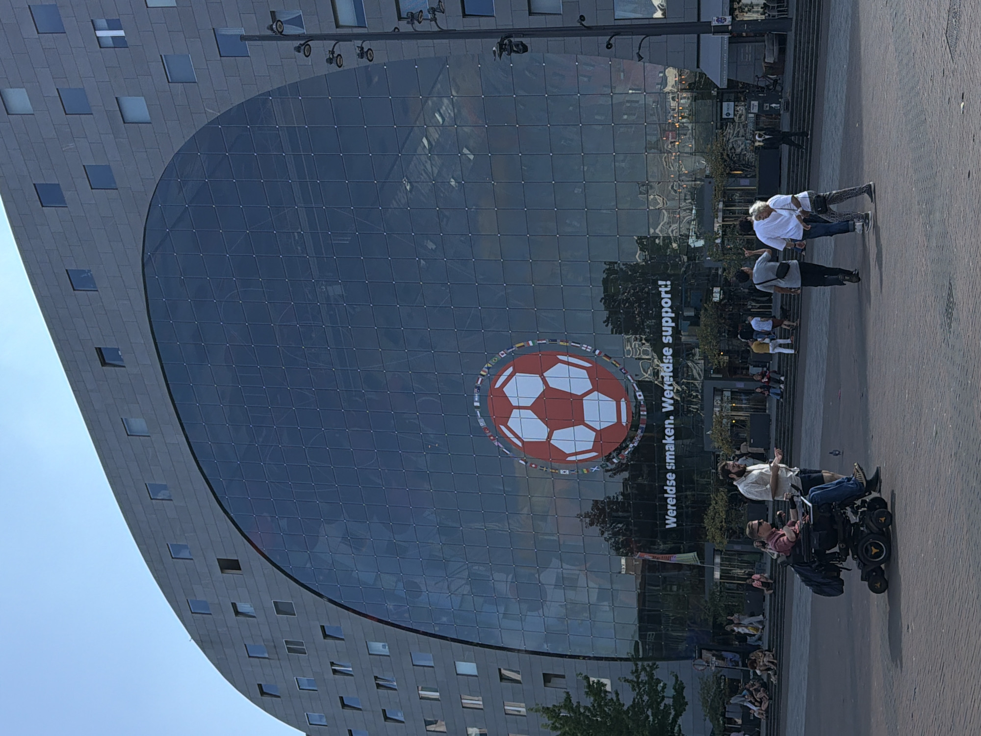 The image depicts the Markthal in Rotterdam, Netherlands, on a sunny day. The massive building, known for its distinctive arching glass facade, dominates the scene. On the glass, a large projection of a red and white soccer ball surrounded by flags of various nations is displayed. Below the projection, text reads "Wereldse smaken. Wereldse support!".
In the foreground, a man in a wheelchair is being pushed by another man. The man in the wheelchair is wearing sunglasses and a plaid shirt, with a laptop on his lap. The other man is wearing a light-colored shirt and shorts. They are walking on a paved square.
Further back, a group of people are seen walking. One man points upwards, while two women walk beside him. In the background, more people are visible walking up a set of stairs and sitting on the steps. Trees and streetlights are also present in the environment. The overall impression is of a busy urban public space.