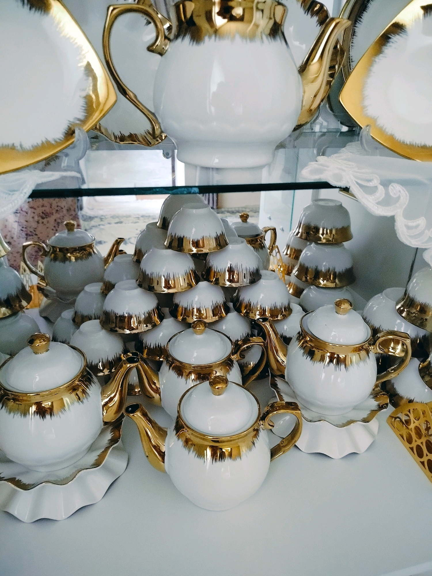 The image displays a collection of white ceramic tea sets with gold accents, likely for sale in a store. The items are arranged on glass shelves. In the foreground, a white teapot with a gold lid and handle is prominent. Behind it, to the left, another similar teapot is partially visible. Several stacks of white ceramic cups with gold rims are visible throughout the image. The overall presentation suggests a retail environment, possibly a china shop or department store, located in the city of Kölkent, Kazakhstan. There are no people present in the image, and no discernible actions or interactions are taking place. The environment appears to be indoors, with artificial lighting.
