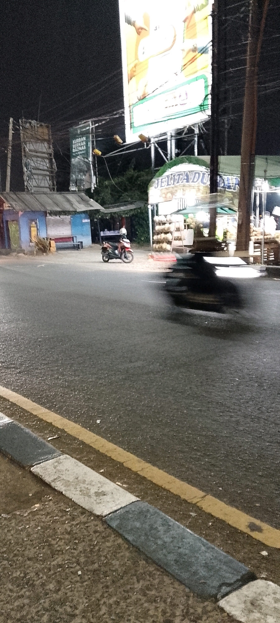 The image depicts a night scene on a street in Depok, Indonesia. A red motorcycle with a white helmet is parked on the side of the road in front of a small, blue and gray structure with a corrugated metal roof. Across the street, a stand displays piles of coconuts under an awning with "JELITA DU" visible on it, suggesting a coconut seller. A large billboard is visible in the background, illuminated by lights. The road surface is wet, indicating recent rain or a damp environment. In the foreground, a section of the sidewalk with gray and white paving stones is visible, with a yellow line marking the edge of the road. A dark, blurred object, likely a moving vehicle, is captured in motion on the right side of the road. The overall atmosphere is that of a typical roadside scene at night in a Southeast Asian city.