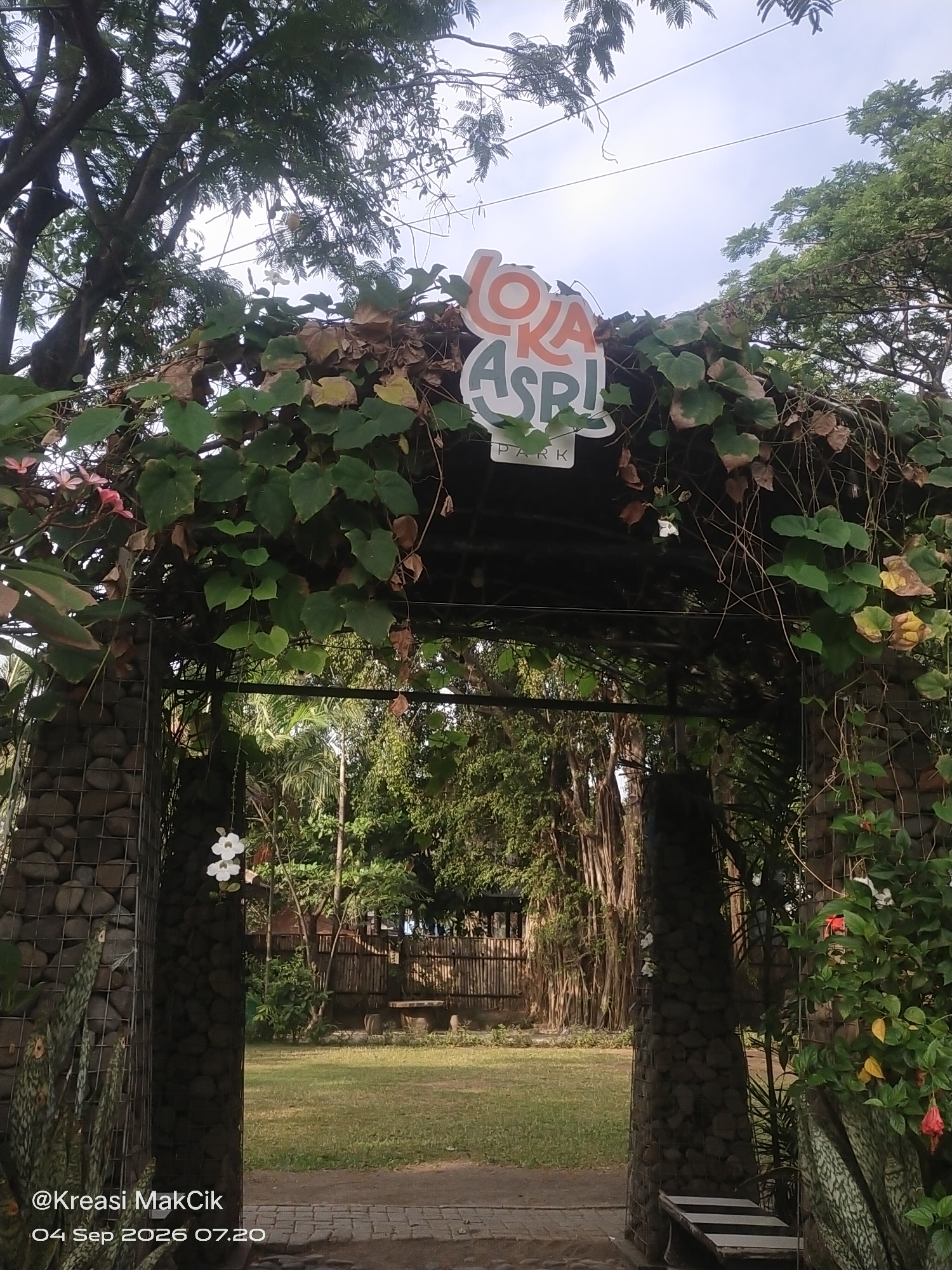 The image captures an entrance archway to "LOKA ASRI PARK" in Suko, Indonesia. The arch is adorned with lush green vines and fallen leaves, framing a view of a grassy area with trees and a bamboo fence in the background. Two stone pillars support the archway, and a bench is visible to the right. The image is a photograph, possibly taken on September 4, 2026, at 7:20 AM, indicated by a watermark in the lower left corner.