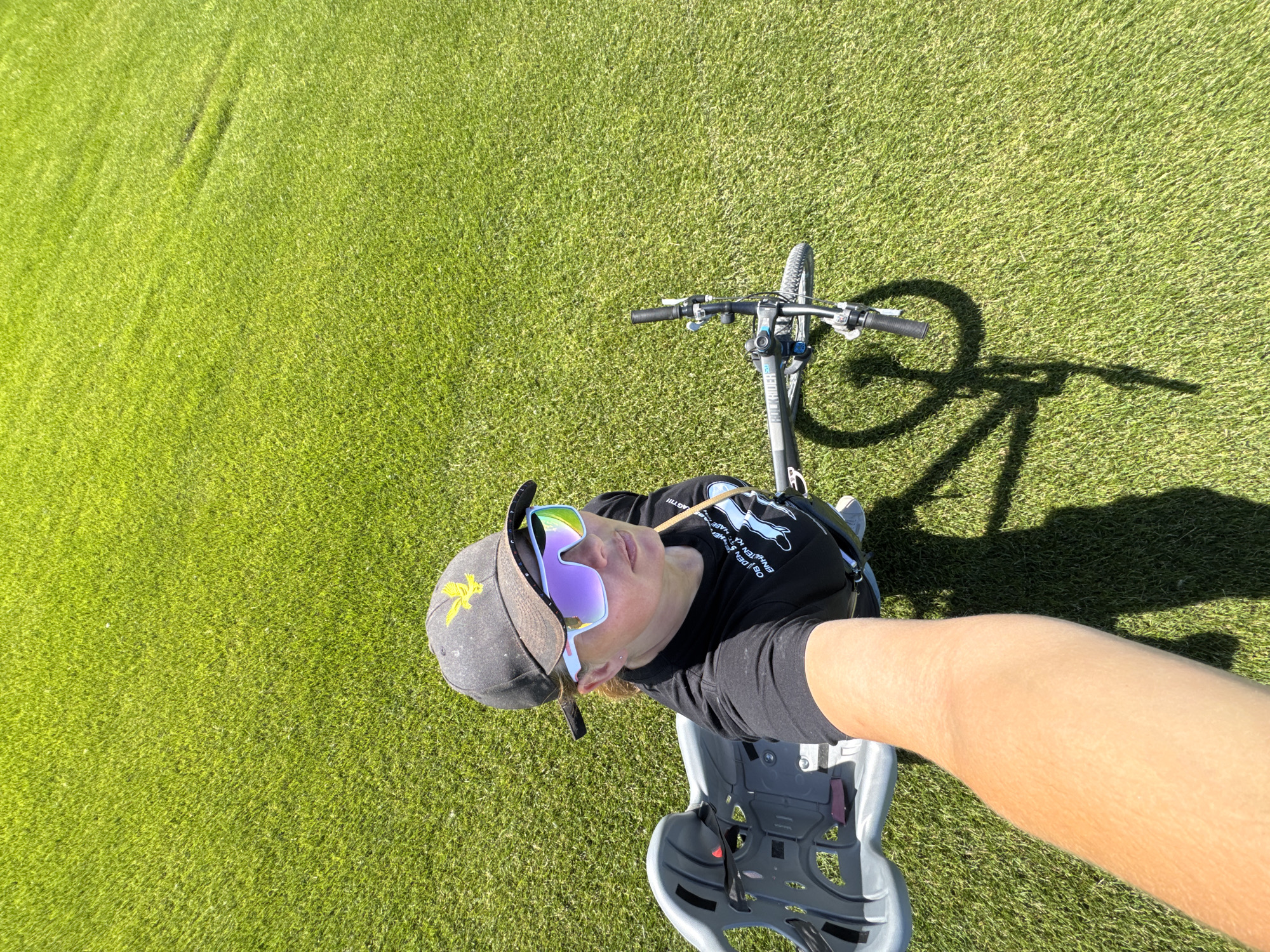 A woman wearing a black t-shirt, a dark baseball cap with a yellow bird emblem, and large, reflective sunglasses is standing on a bright green, well-manicured lawn, likely a park or golf course. She is holding a selfie stick, indicating she is taking a photo of herself. Next to her is a bicycle with a child seat attached. The bicycle is grey and black, and its handlebars are visible. The woman's left arm and hand are extended towards the camera, holding the selfie stick. The background consists entirely of the lush green grass, suggesting an outdoor setting on a sunny day. The scene appears to be taking place in the city of Kempelevo, Russia.