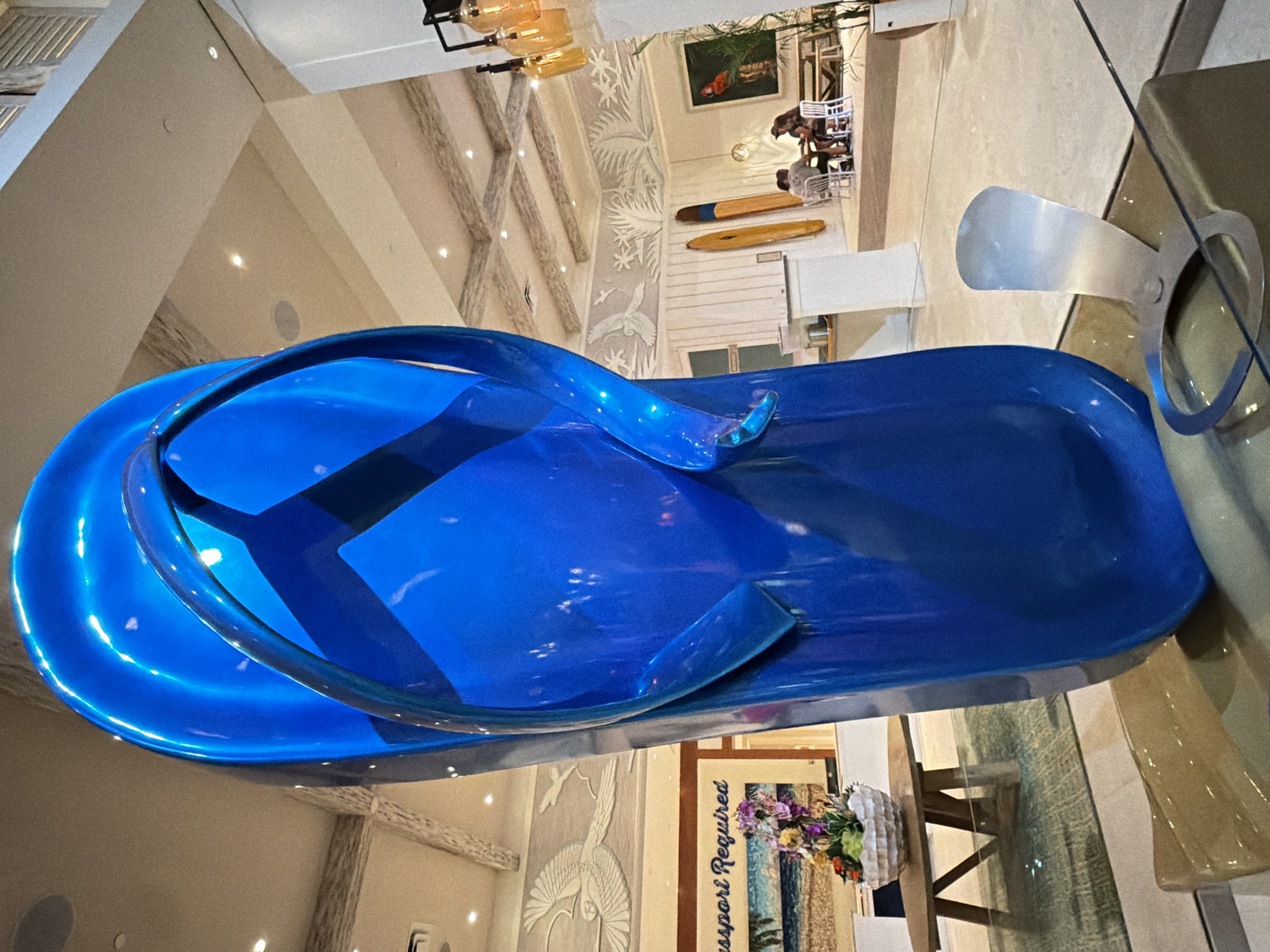 This is a wide shot of a large, shiny blue flip-flop sculpture in a brightly lit, modern lobby. The sculpture is the primary focus, dominating the foreground and mid-ground. In the background, there is a seating area where two people are seated at a table. There are also surfboards displayed on the wall, a painting of a parrot, and decorative wall art depicting birds and palm fronds. The ceiling has a coffered design with recessed lighting. The scene appears to be located in a hotel or resort lobby, possibly in Hollywood, Florida, given the tropical-themed decor.