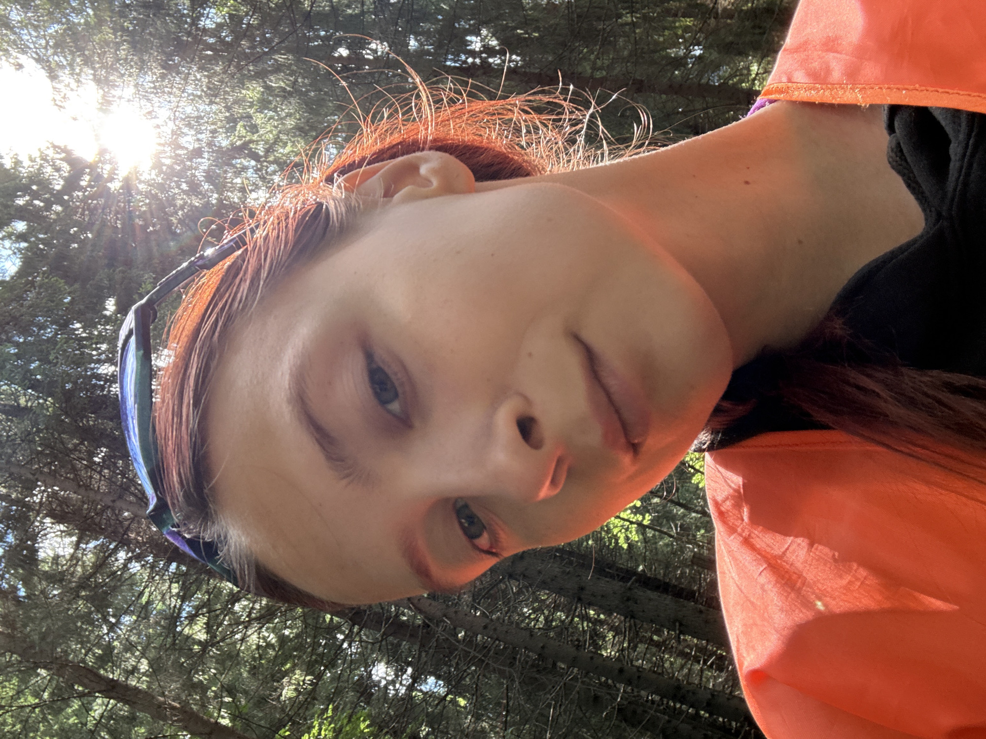 A young woman with reddish-brown hair, wearing sunglasses on her head and an orange vest over a black top, poses for a selfie in a sun-dappled forest. The background consists of tall, dense evergreen trees. The sun is bright and appears to be setting or rising, casting strong highlights and lens flare across the image. The woman's expression is relaxed, with a slight smile. The location is not identified as Сергино, Russia, but the environment suggests an outdoor, natural setting, possibly a park or wilderness area.