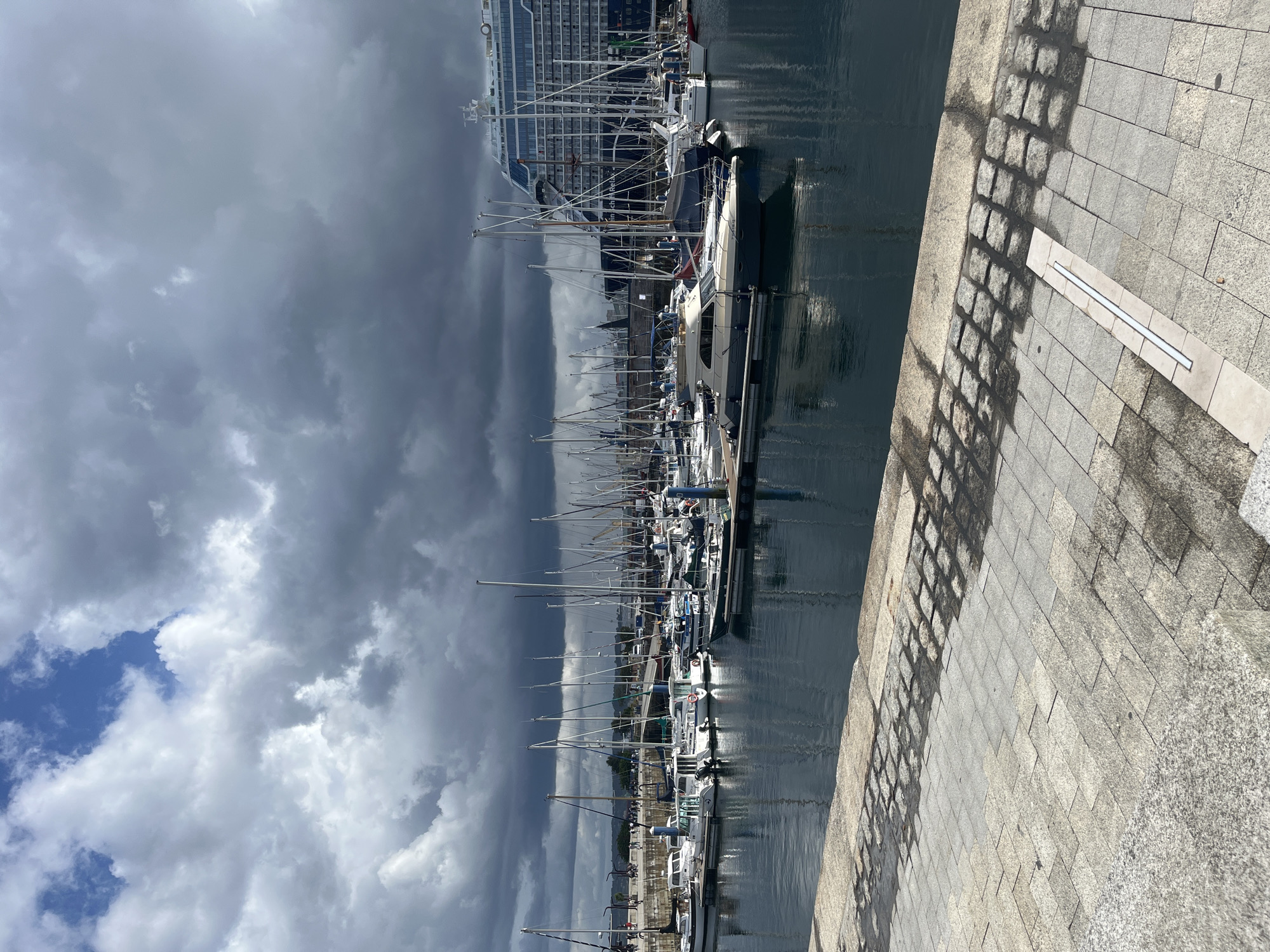 The image shows a harbor in A Coruña, Spain, filled with many yachts docked along a quay. A large cruise ship is visible in the background. The sky is overcast with a mix of dark, heavy clouds and patches of blue sky. The quay is made of light-colored stone tiles. The water in the harbor reflects the clouds and boats. Shadows from a railing or fence are cast onto the quay. There are no people visible in the image.