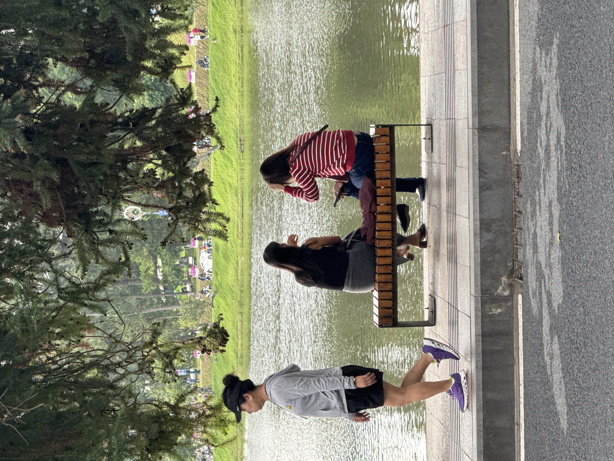 This image depicts a serene daytime scene at a lakeside park in Da Lat, Vietnam. Three women are observed enjoying the peaceful environment.
In the foreground, a woman stands facing to the left, dressed in a grey hoodie, black shorts, a black visor, and distinctive purple and white athletic shoes. Her hair is tied back, and she appears to be observing the surroundings.
To her right, two other women are seated on a wooden bench positioned along a paved walkway that runs beside a calm body of water. The woman closer to the viewer is wearing a black top and a grey skirt, gazing out towards the shimmering lake. Further down the bench, another woman in a red and white striped long-sleeved shirt and blue jeans leans forward, seemingly preoccupied with something in her hands.
The sparkling water, possibly a lake or pond, occupies the mid-ground, reflecting sunlight. Beyond the water, a lush green bank transitions into a line of trees. In the distant background, more dense foliage and subtle hints of structures, including what resembles a Ferris wheel, suggest a larger recreational area. Large coniferous trees with prominent branches frame the left side of the composition. An asphalt path runs parallel to the tiled walkway on the right. The overall atmosphere conveys relaxation and tranquility. No visible text is present.
